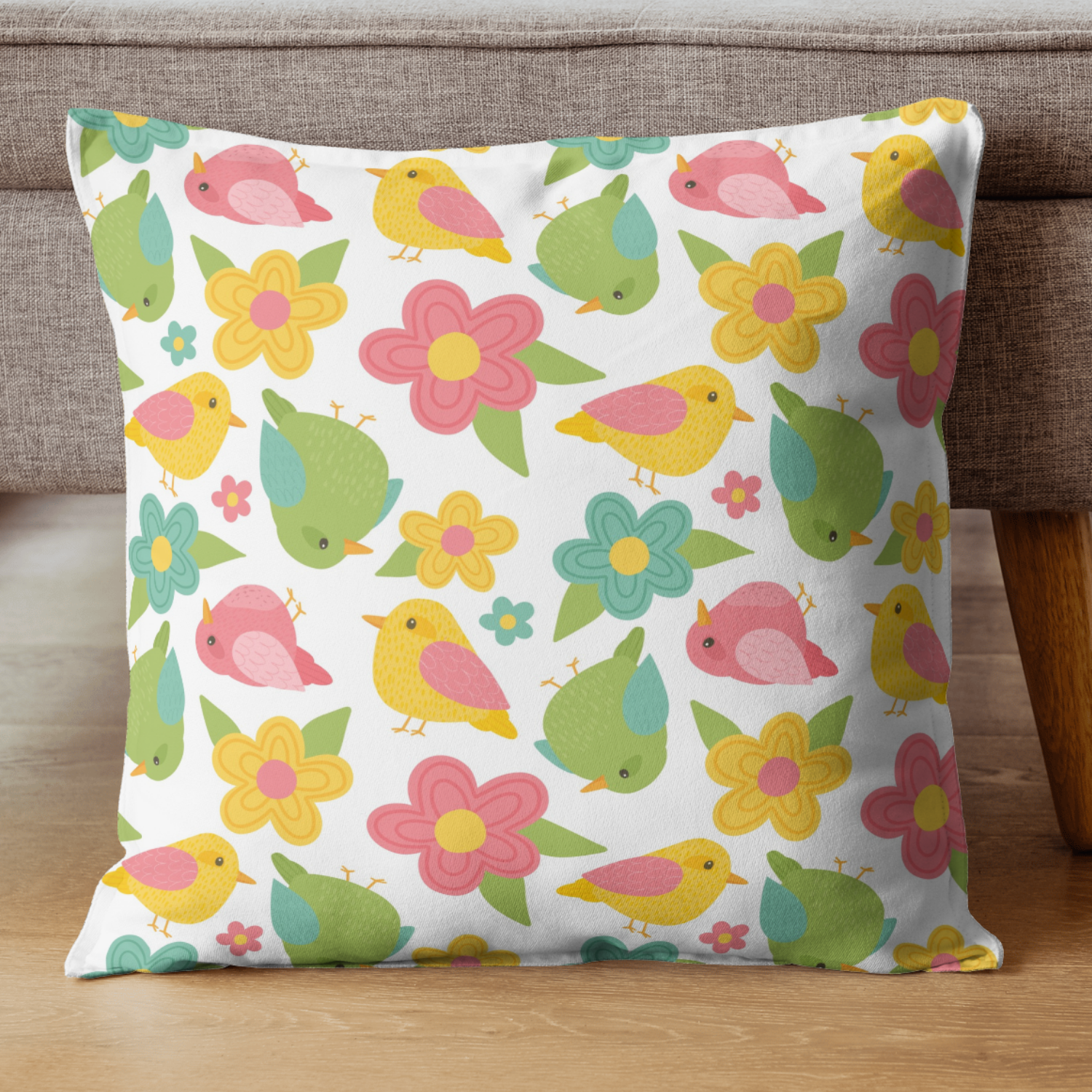 A decorative pillow with a colorful pattern of yellow, pink, green, and blue birds and flowers on a white background, placed on a wooden surface against a brown cushion.