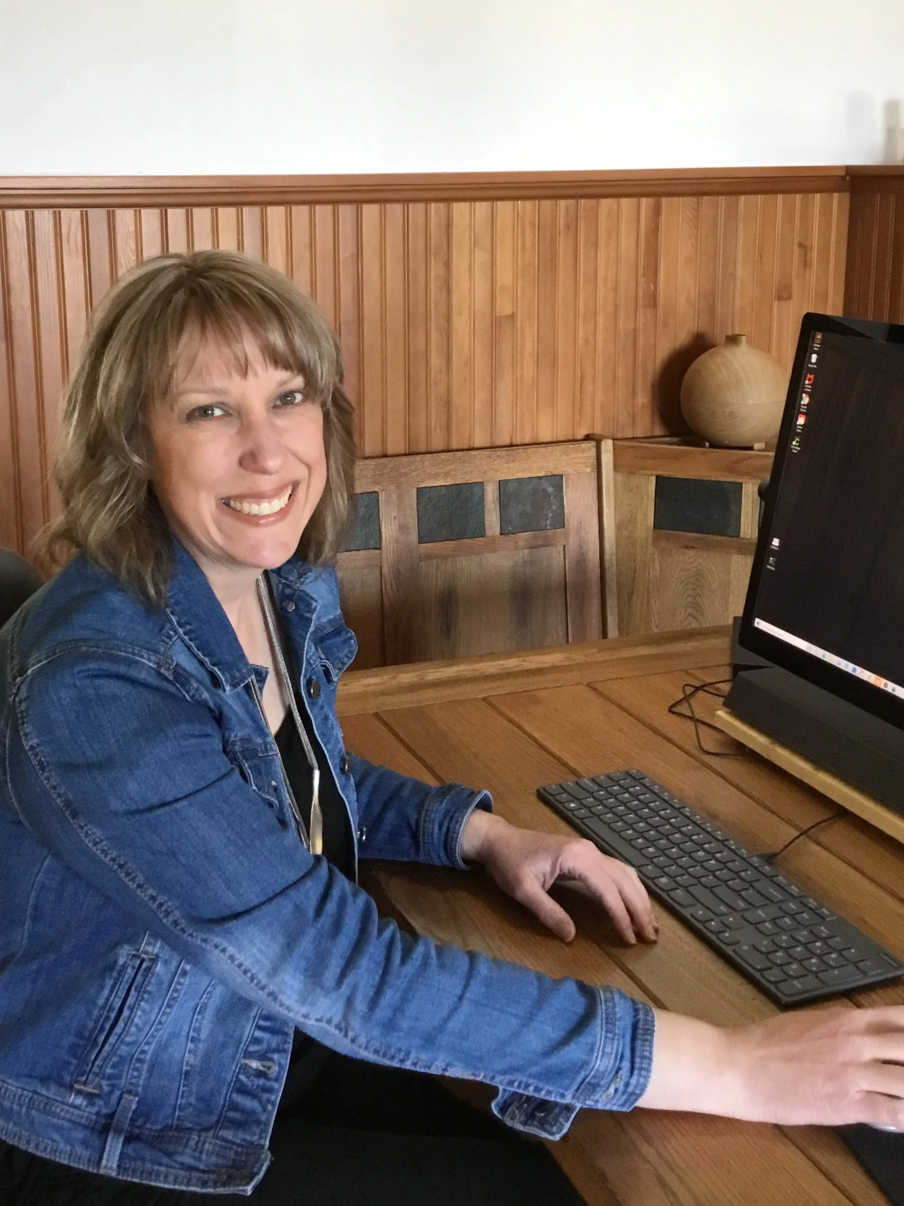 Woman sitting at a wooden table in front of a computer, smiling, wearing a blue denim jacket, with a wooden wall background and a decorative pottery vase on the side.