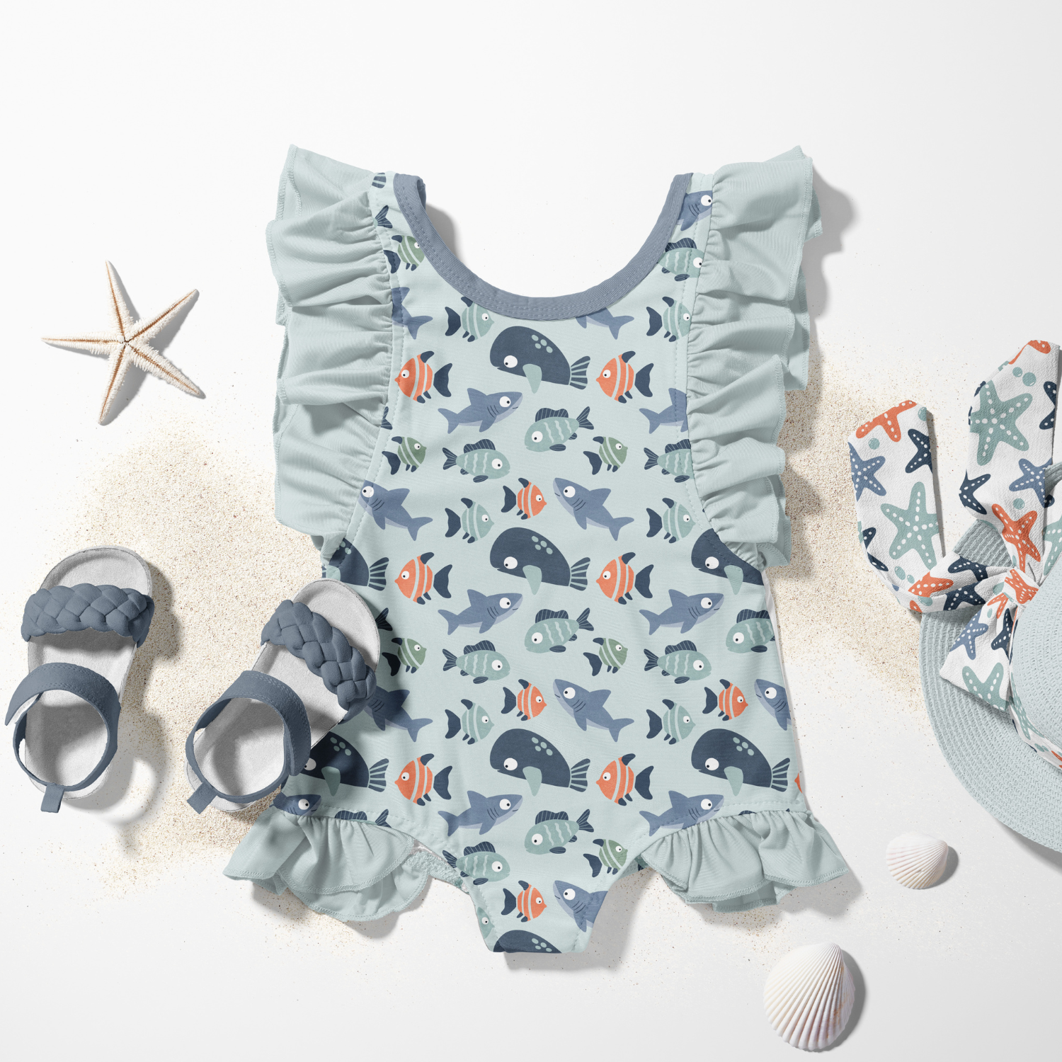Baby swimsuit with fish pattern, matching starfish and seashells, and baby sandals on a sandy surface.