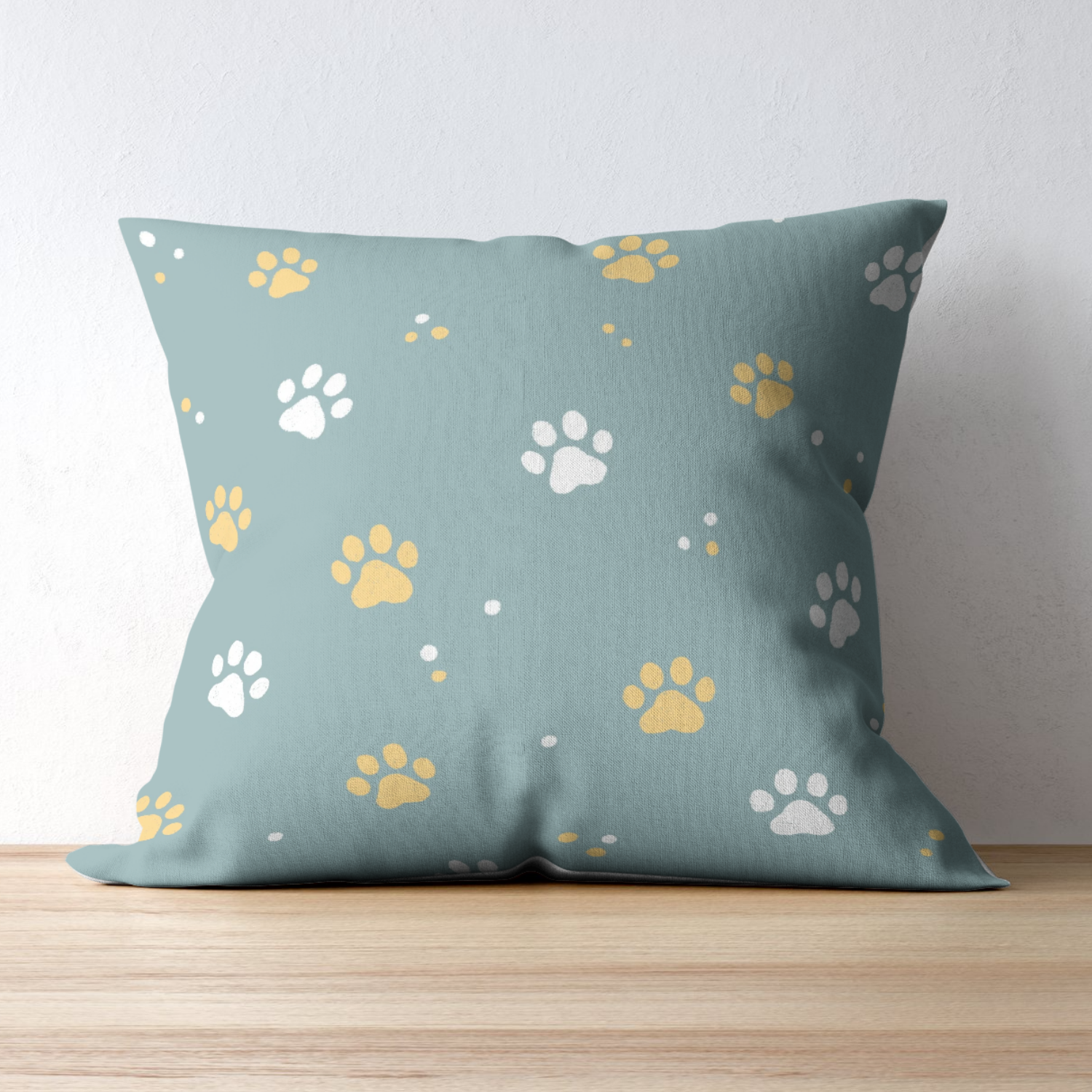 Light blue pillow with white and yellow paw print patterns resting on a wooden surface against a white wall.