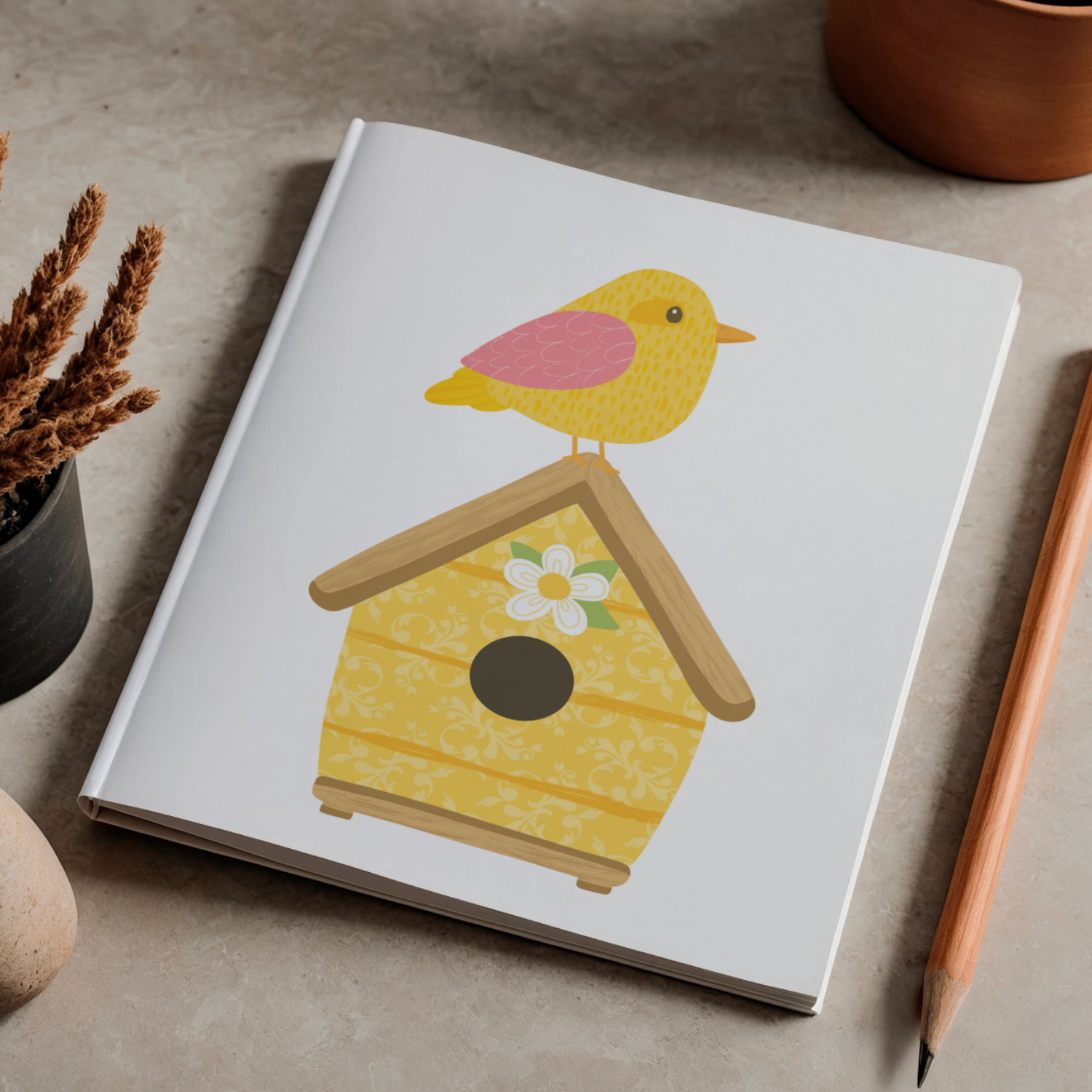 Notebook with a birdhouse and a yellow bird illustration on its cover, placed on a tabletop next to a pencil and decorative plants.