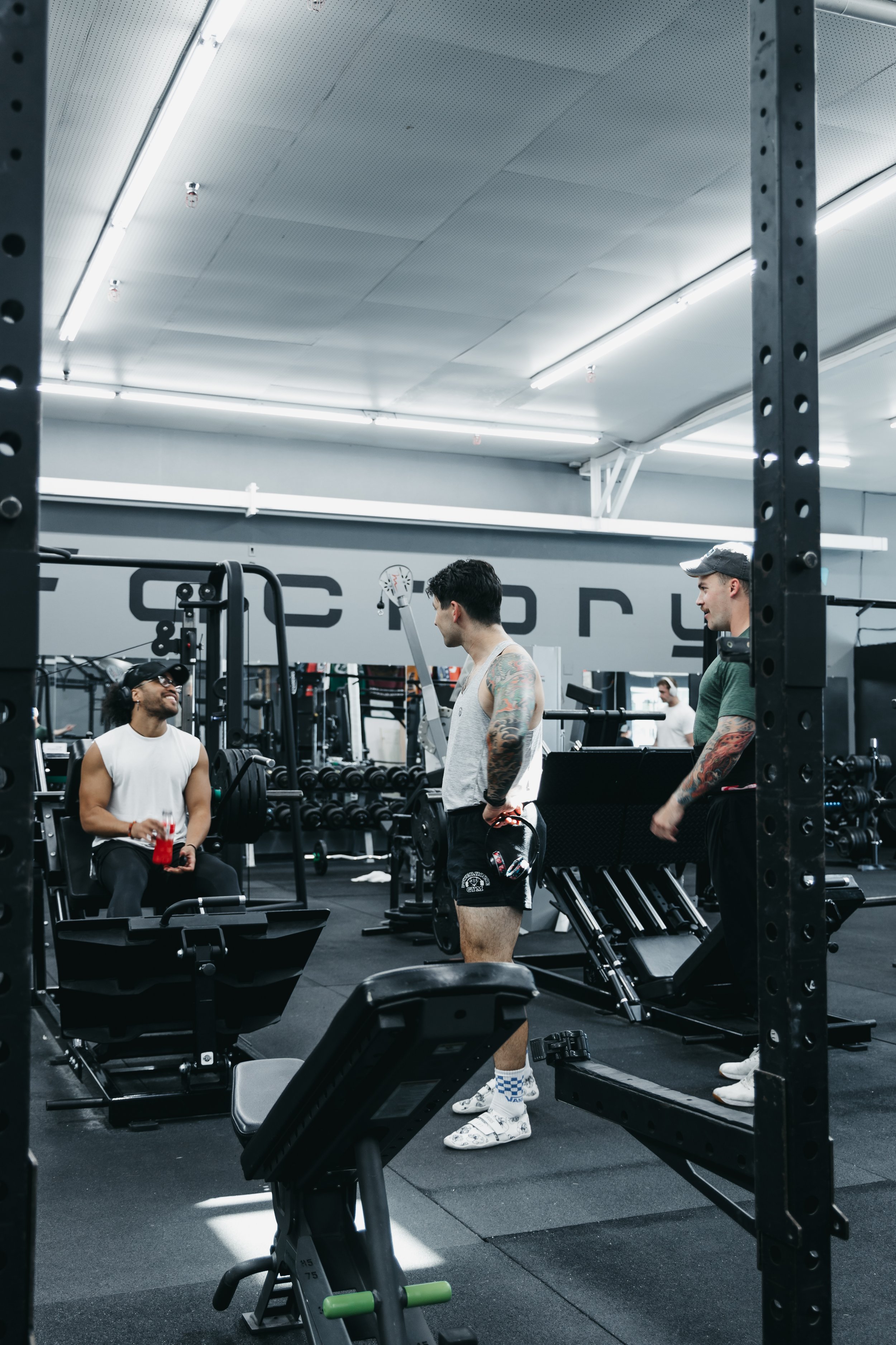 Inside a gym, three men are talking and smiling near workout equipment. One man sitting on a bench is wearing a white tank top, black pants, sunglasses, and a baseball cap, holding a red water bottle. Two other men are standing, one with a gray tank top and tattoos, and the other with a green shirt and gray cap. The gym has black flooring, weight racks, and bright overhead lighting.