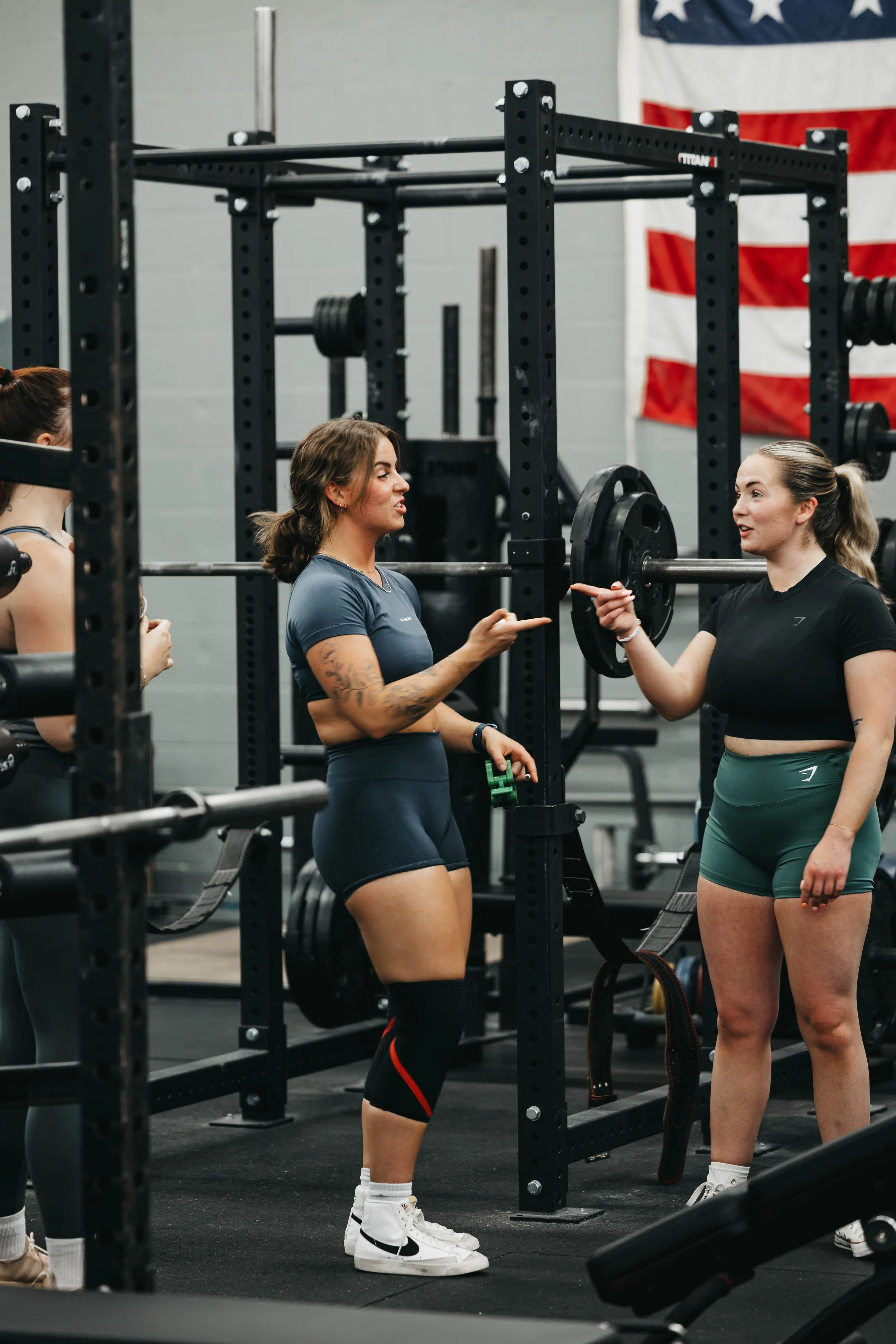 Two women at a gym engaged in conversation, standing near a squat rack with weights, with an American flag in the background.