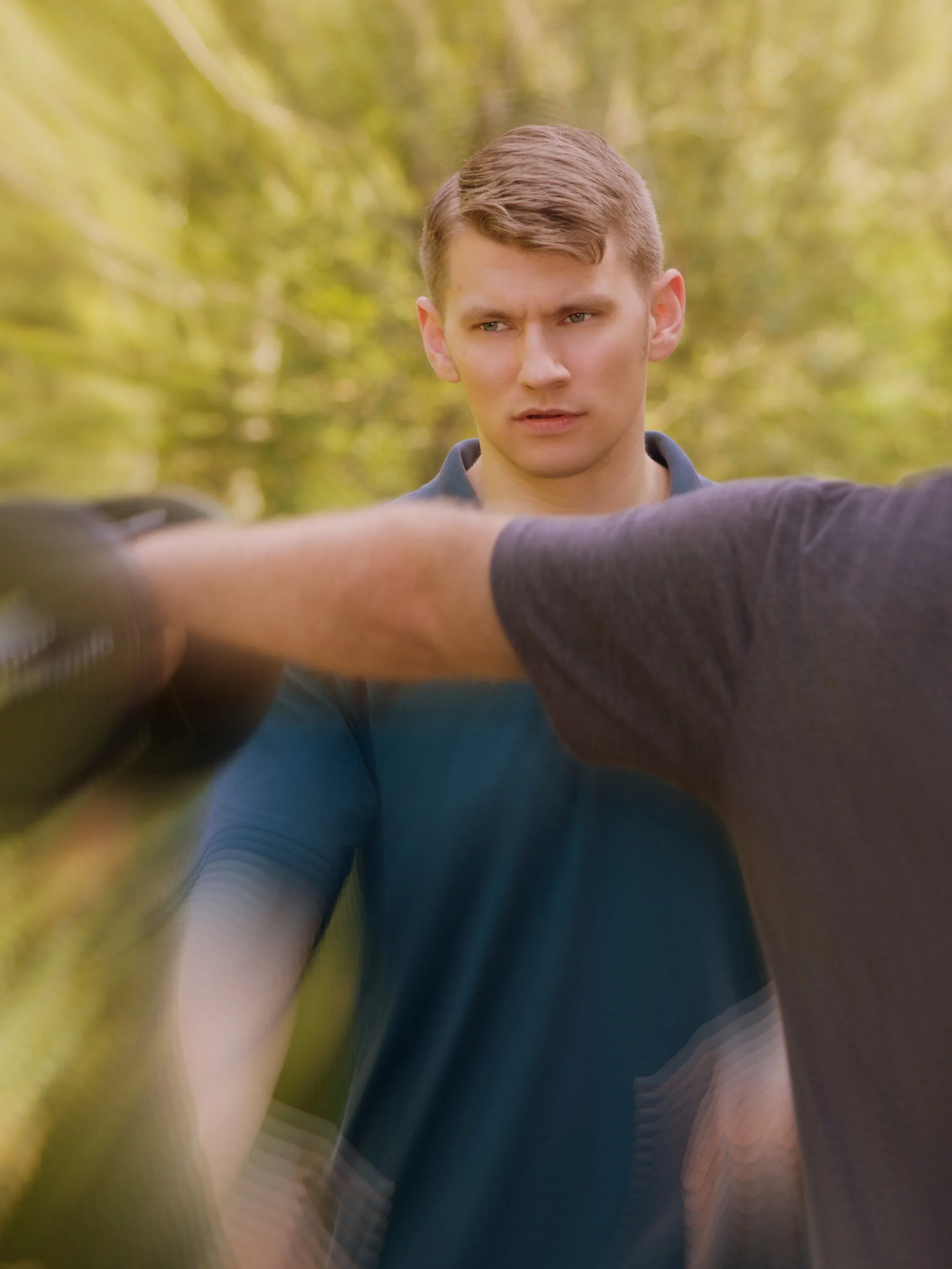 Close-up of a young man with short brown hair and blue eyes, wearing a dark blue shirt, standing outdoors with blurry trees and sunlight in the background.