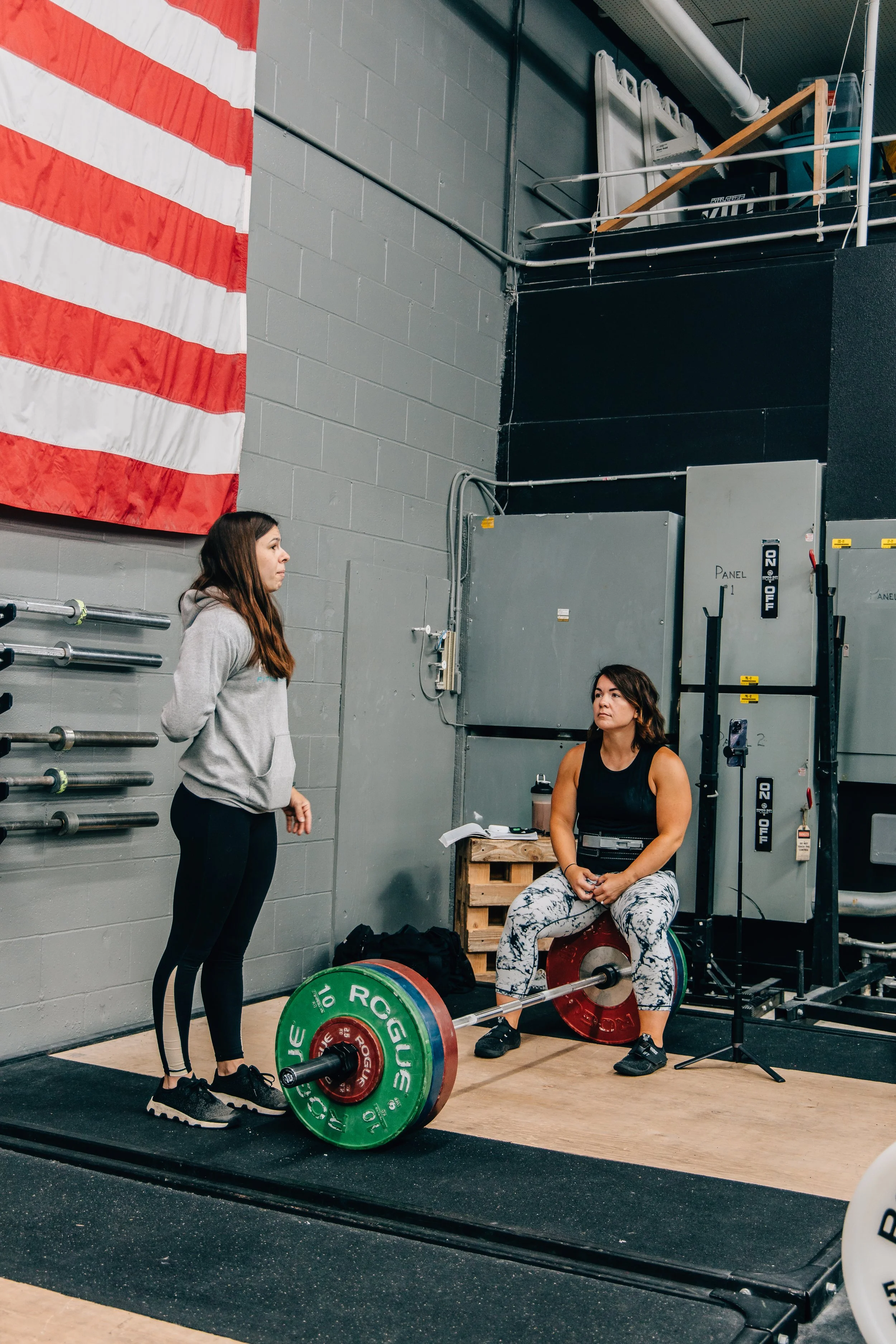 Two women in a gym, one standing and one sitting, with weights and gym equipment around them. The American flag is hanging on the wall.