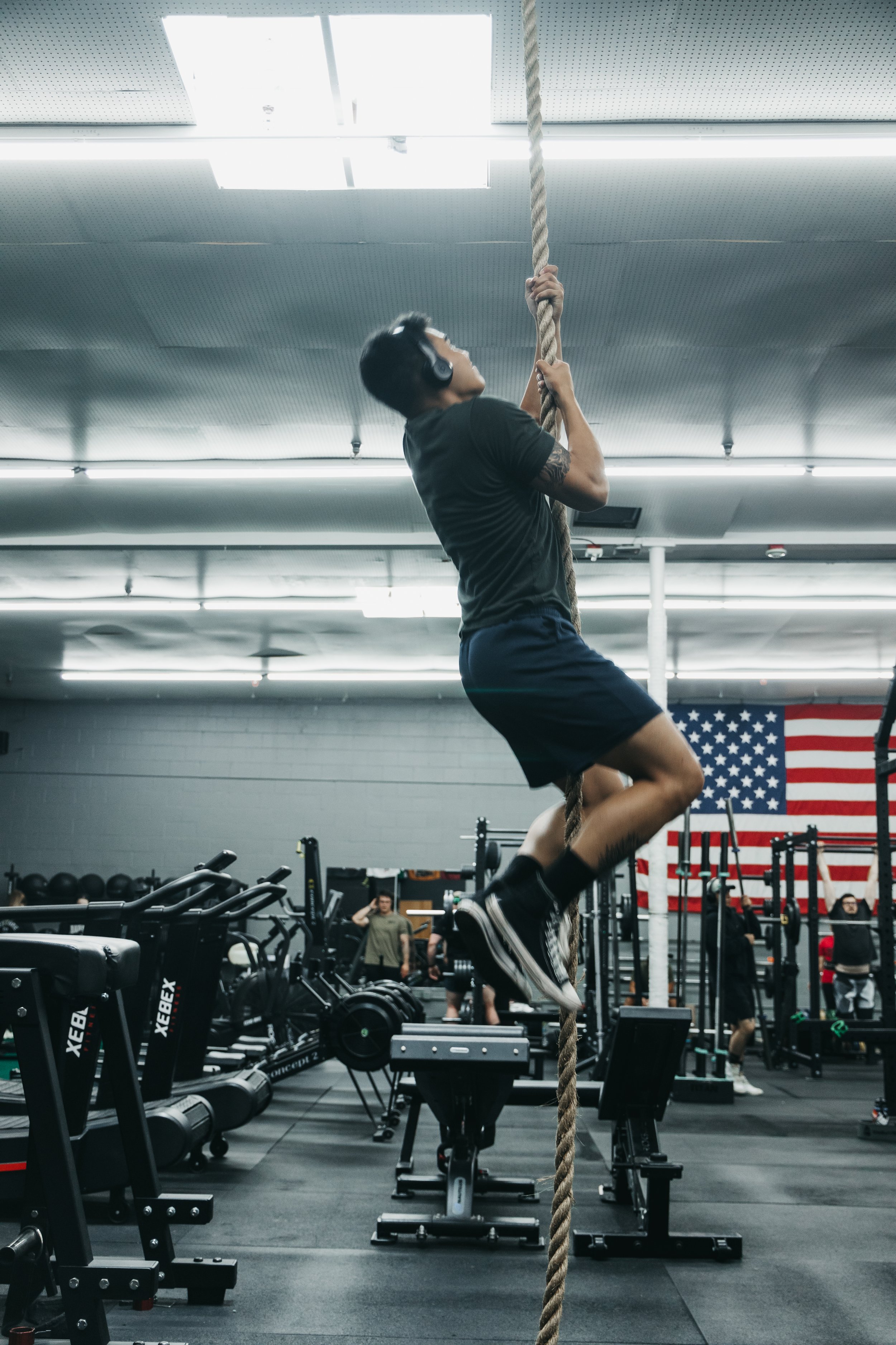 A person wearing headphones climbing a thick rope in a gym. The gym has black equipment, American flag hanging on the wall, and several people in the background.