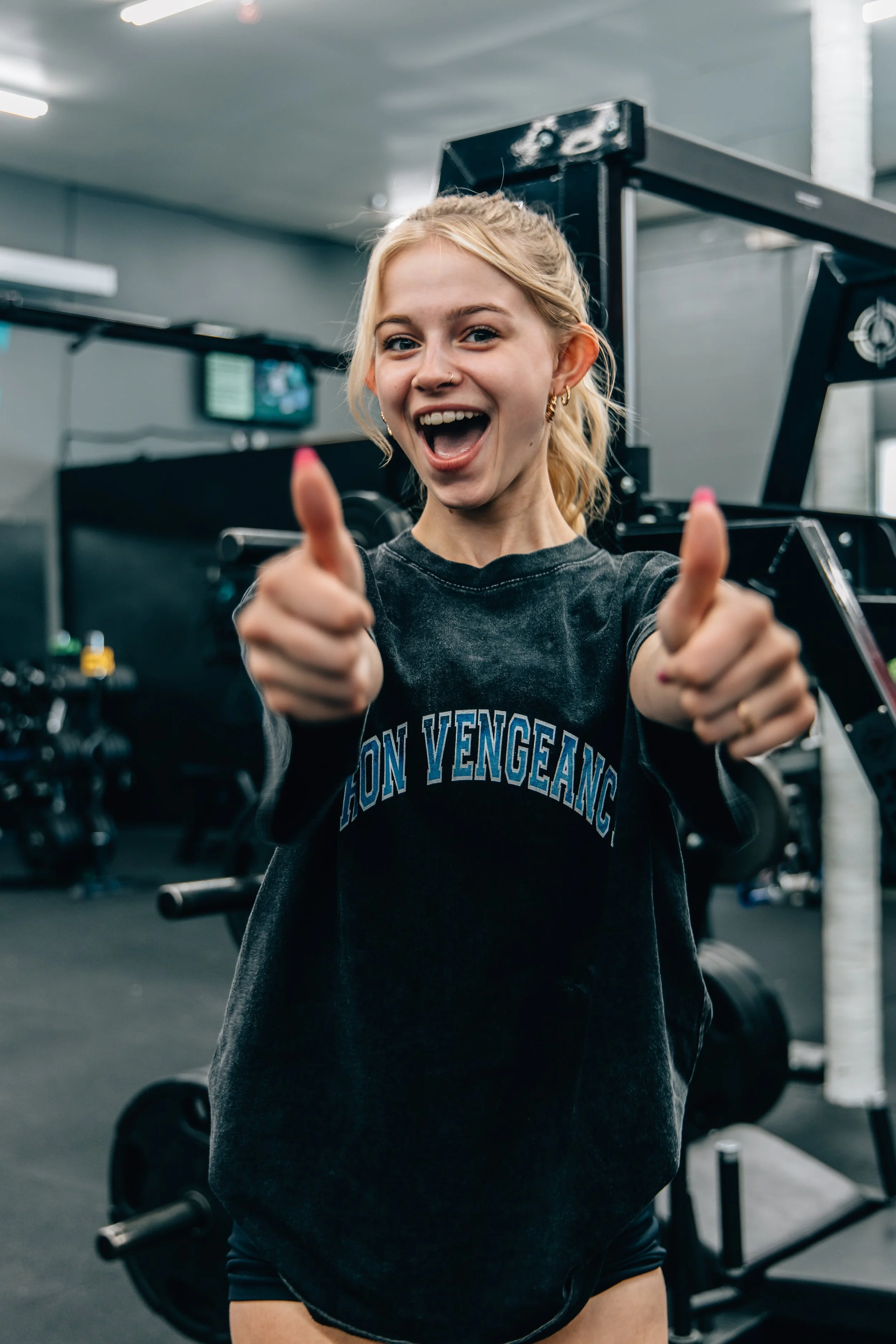 Young woman with blonde hair tied back, giving a thumbs-up, smiling, in a gym with workout equipment in the background.