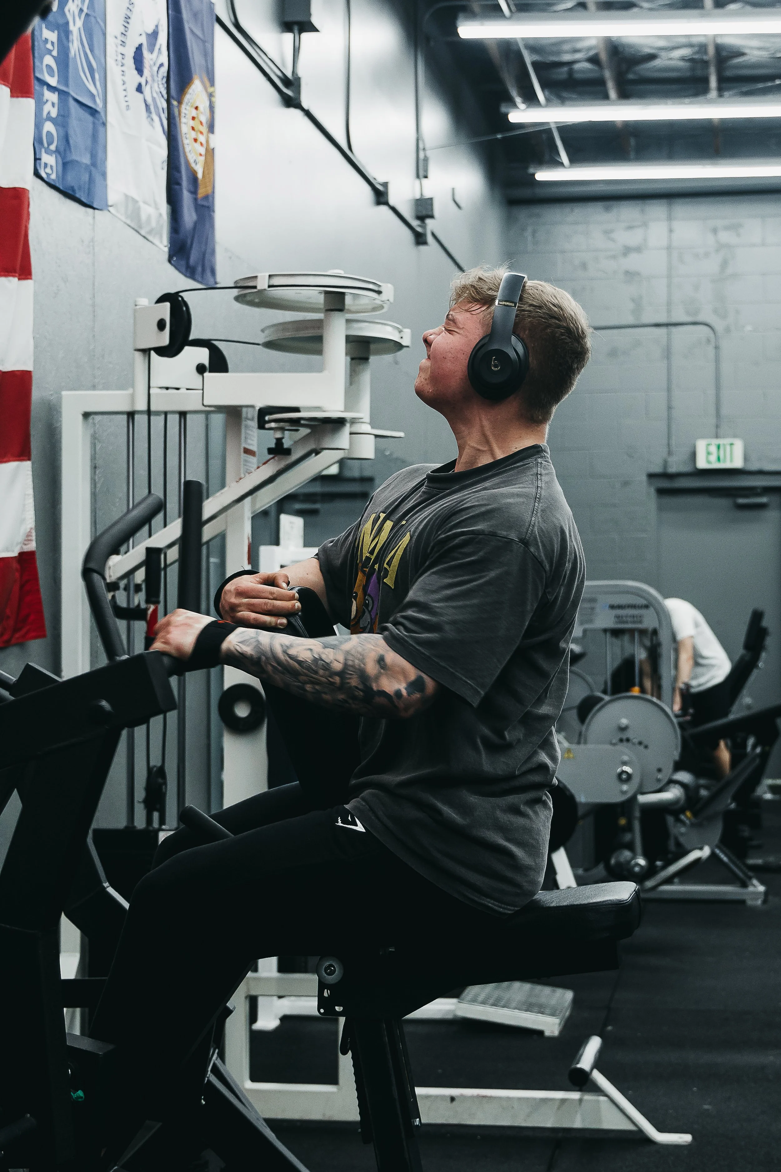 A man with tattoos on his arm wearing headphones and a T-shirt, performing a row on a piece of equipment in a gym, exerting himself with his eyes closed.
