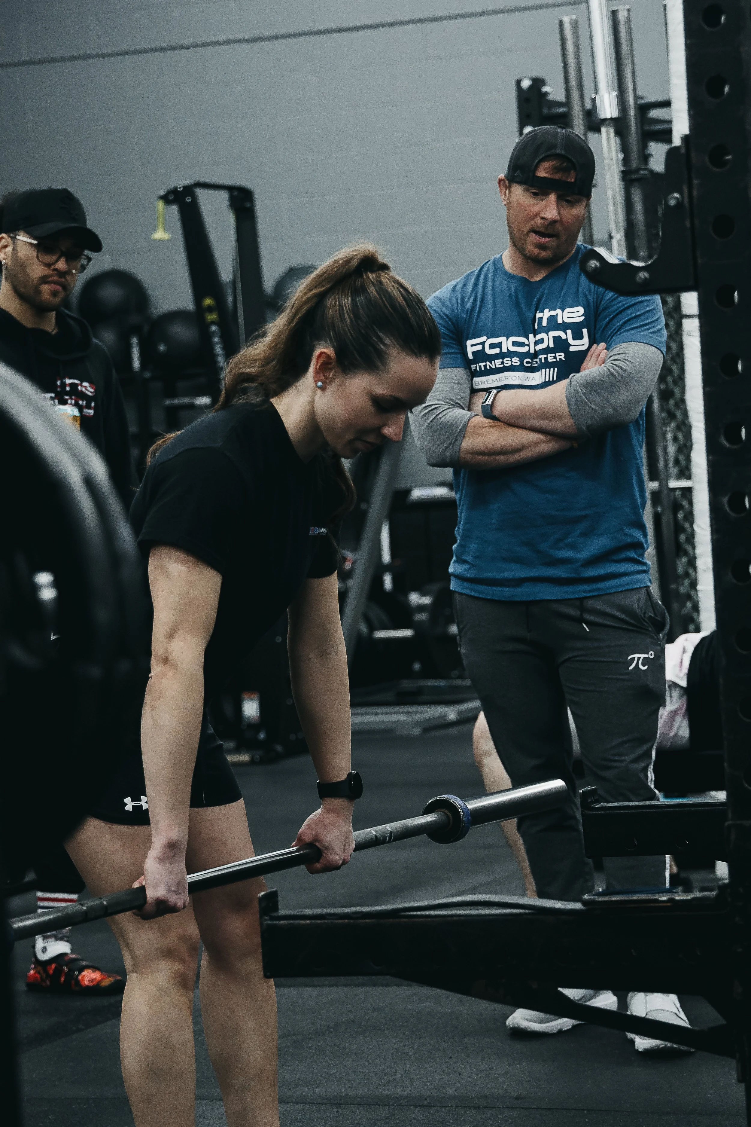 A woman in black athletic wear lifting a barbell at a gym while being coached by a man in a blue shirt and black cap. Other gym equipment can be seen in the background.