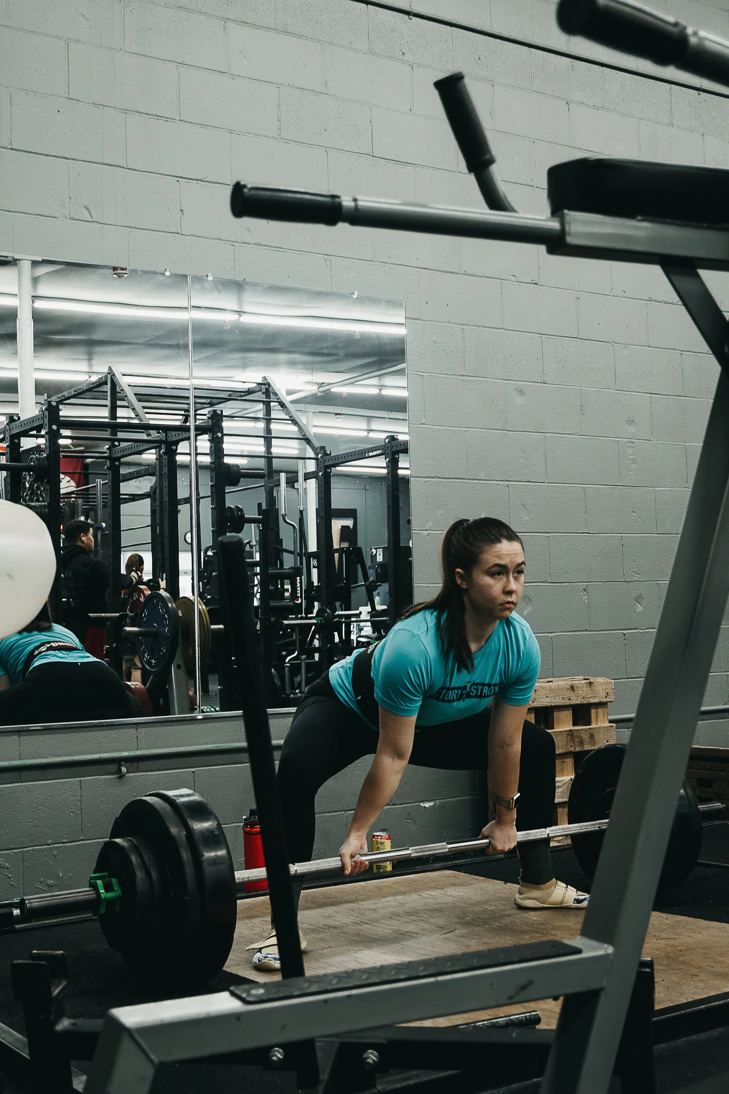 A woman in a blue shirt deadlifitng in a gym, with gym equipment and a mirror visible in the background.