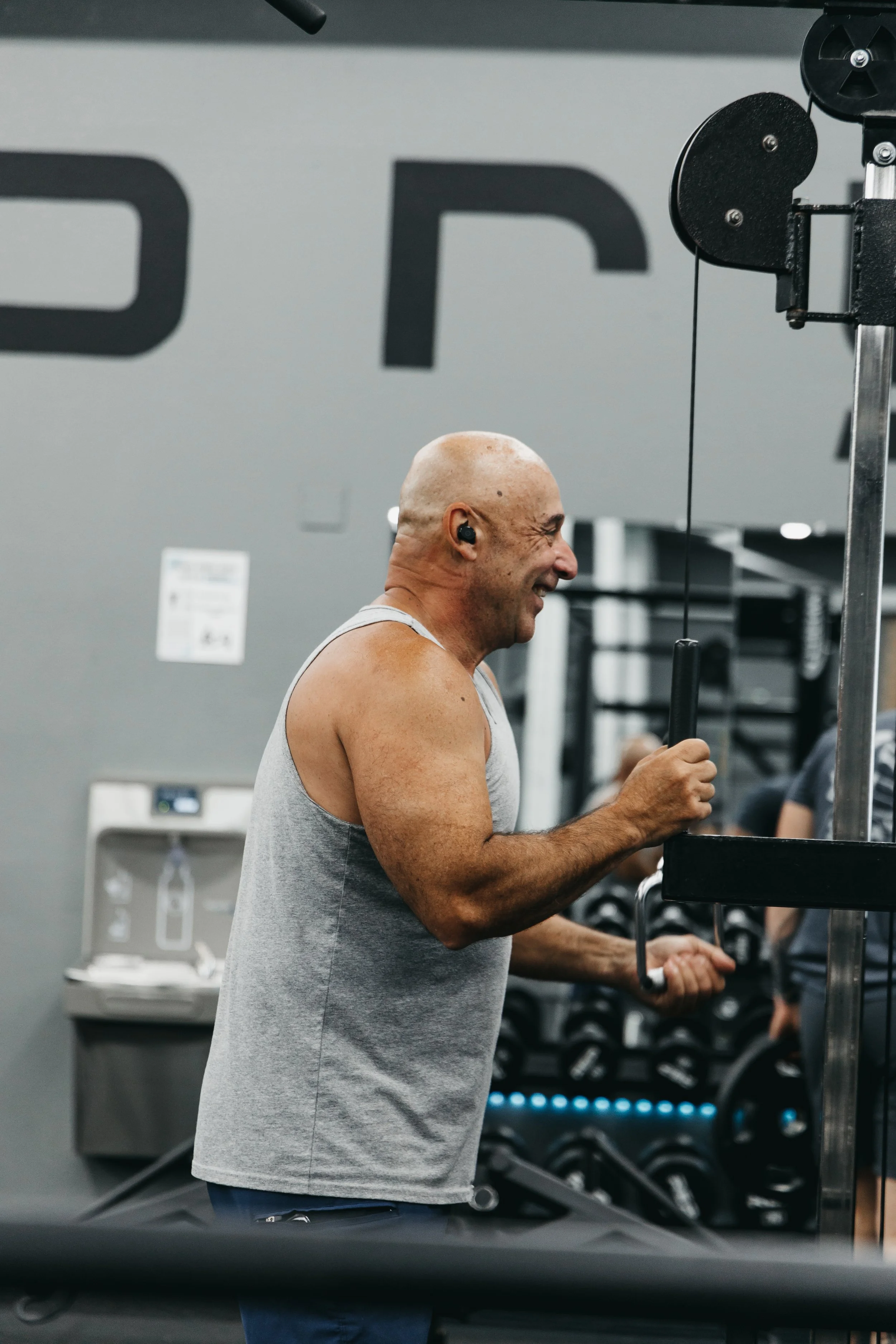 A bald man smiling and wearing a gray sleeveless shirt, exercising at the gym on a machine.