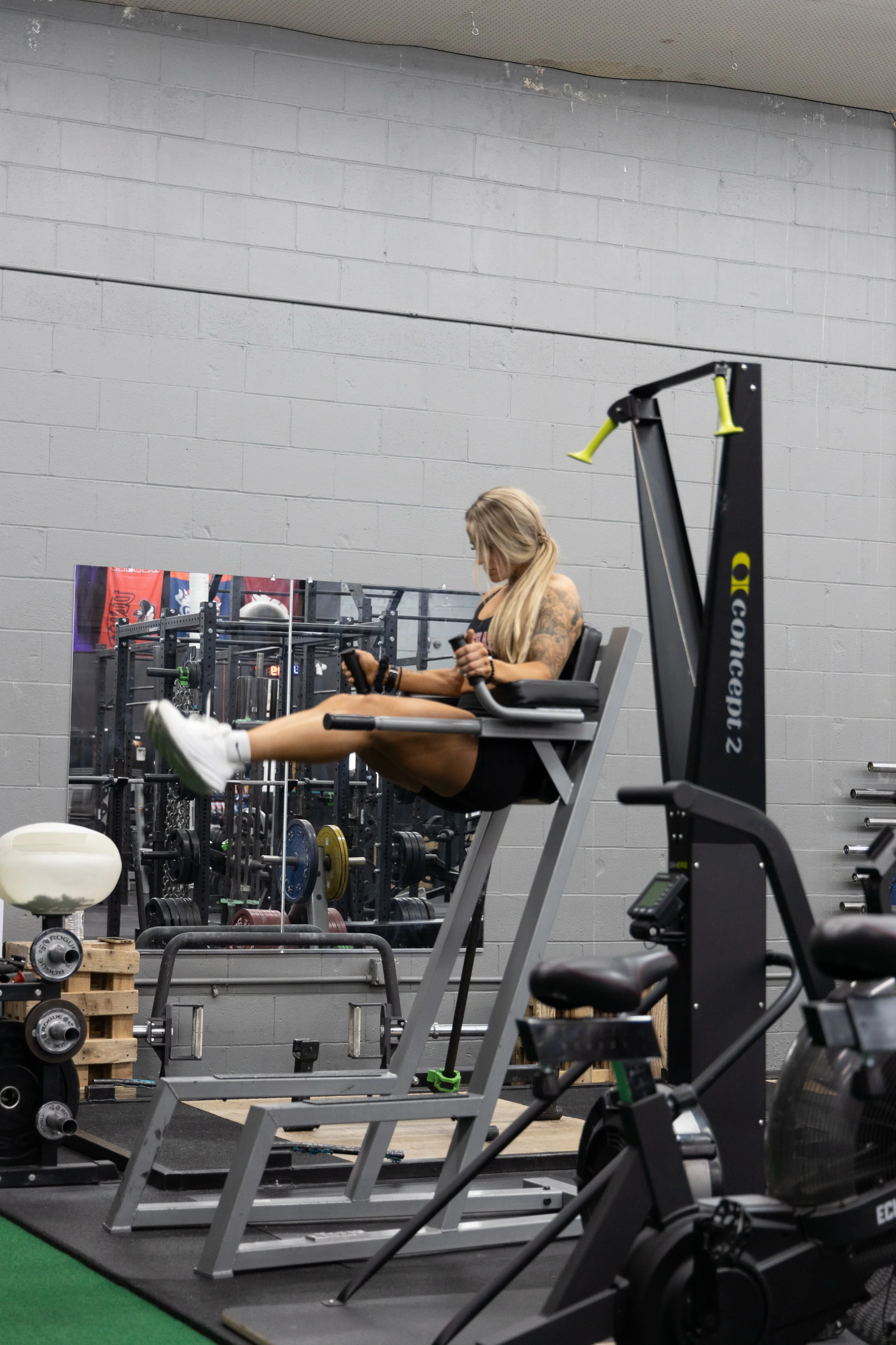 A woman with blonde hair performing leg raises in a gym, wearing black workout clothes and, sneakers, with gym equipment and a wall mirror in the background.