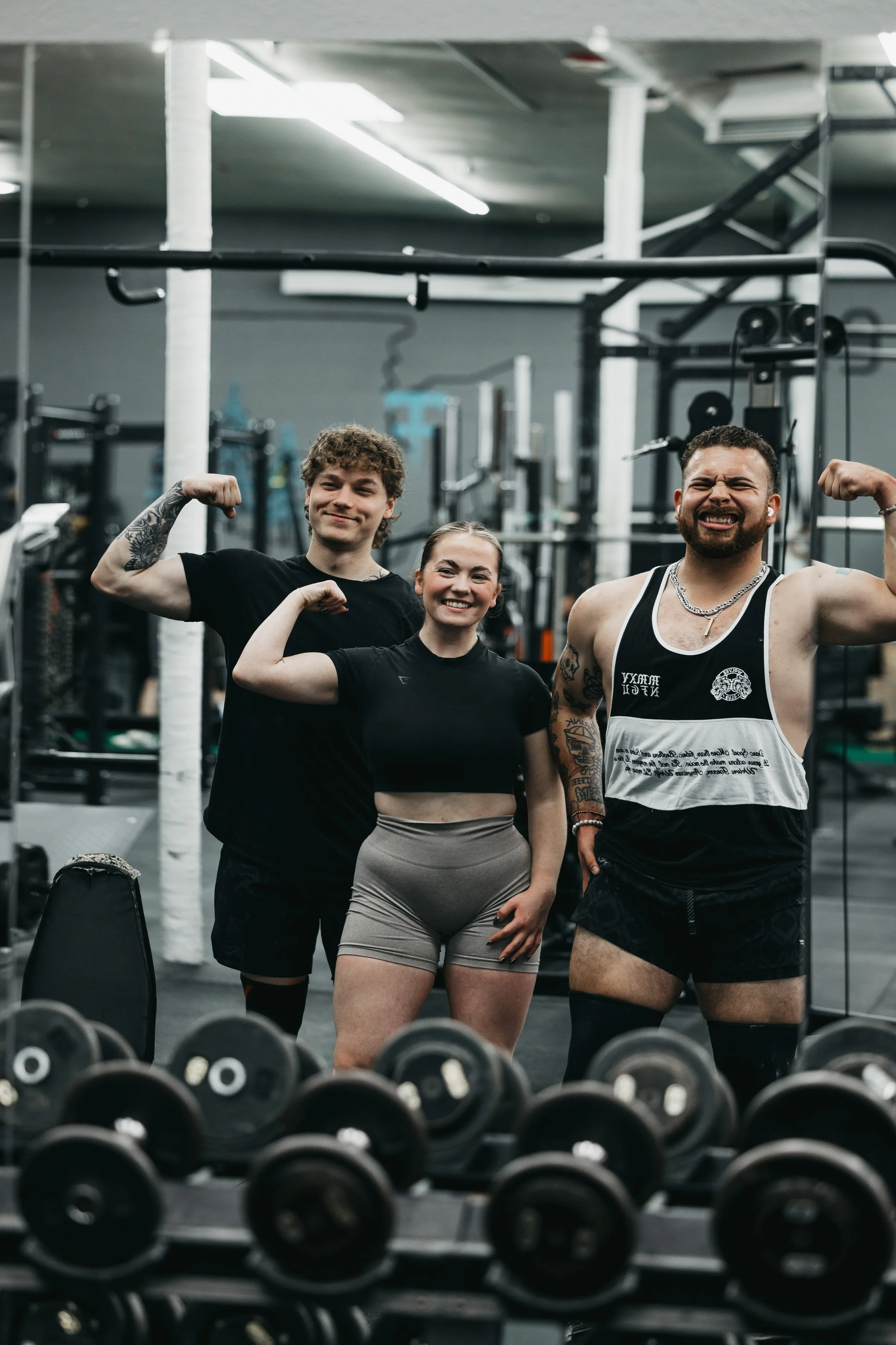 Three individuals in a gym flexing their muscles. Two men and a woman, all smiling.