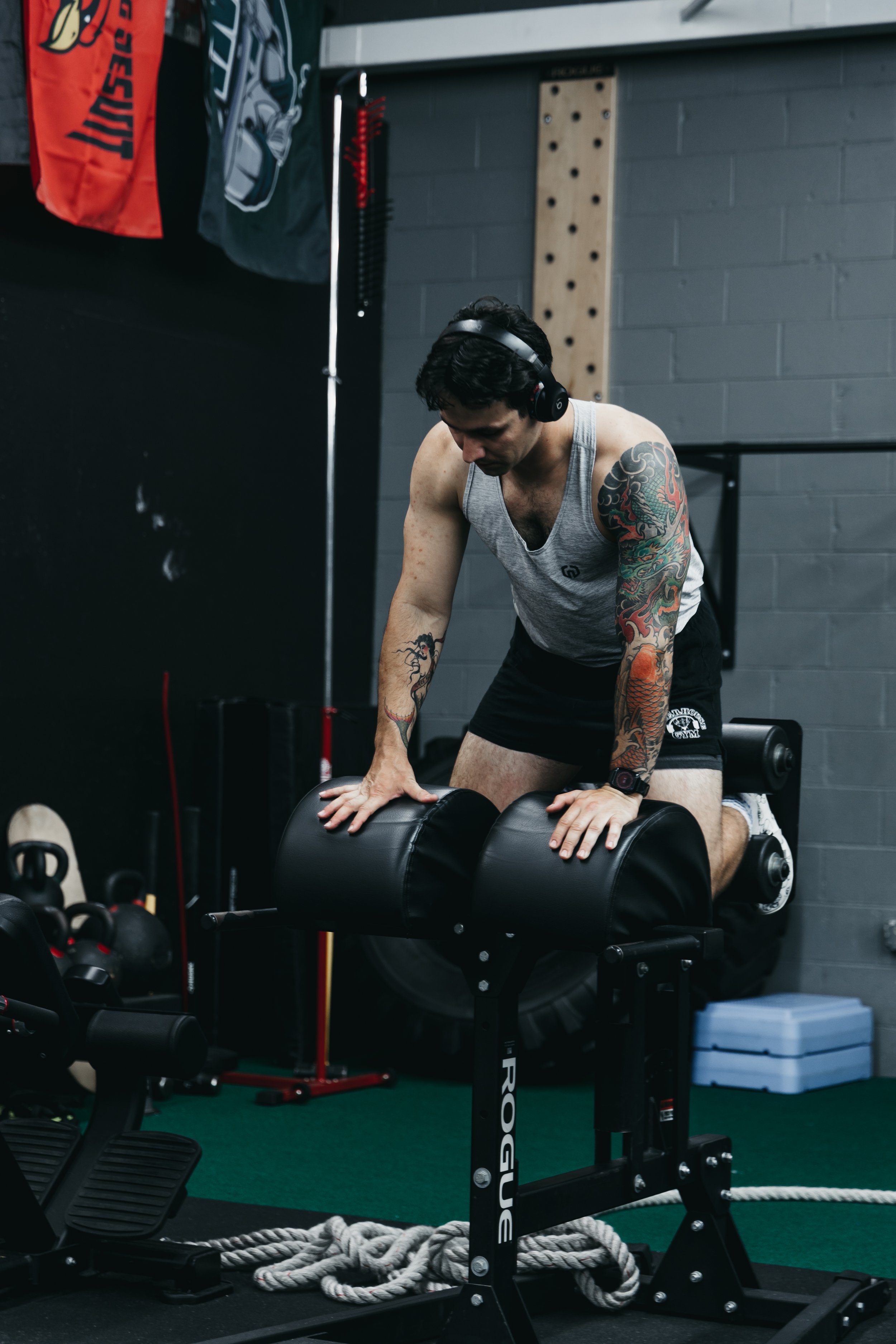 Man resting after doing glute hamstring raises in a gym, wearing a gray tank top, black shorts, headphones, and a watch.