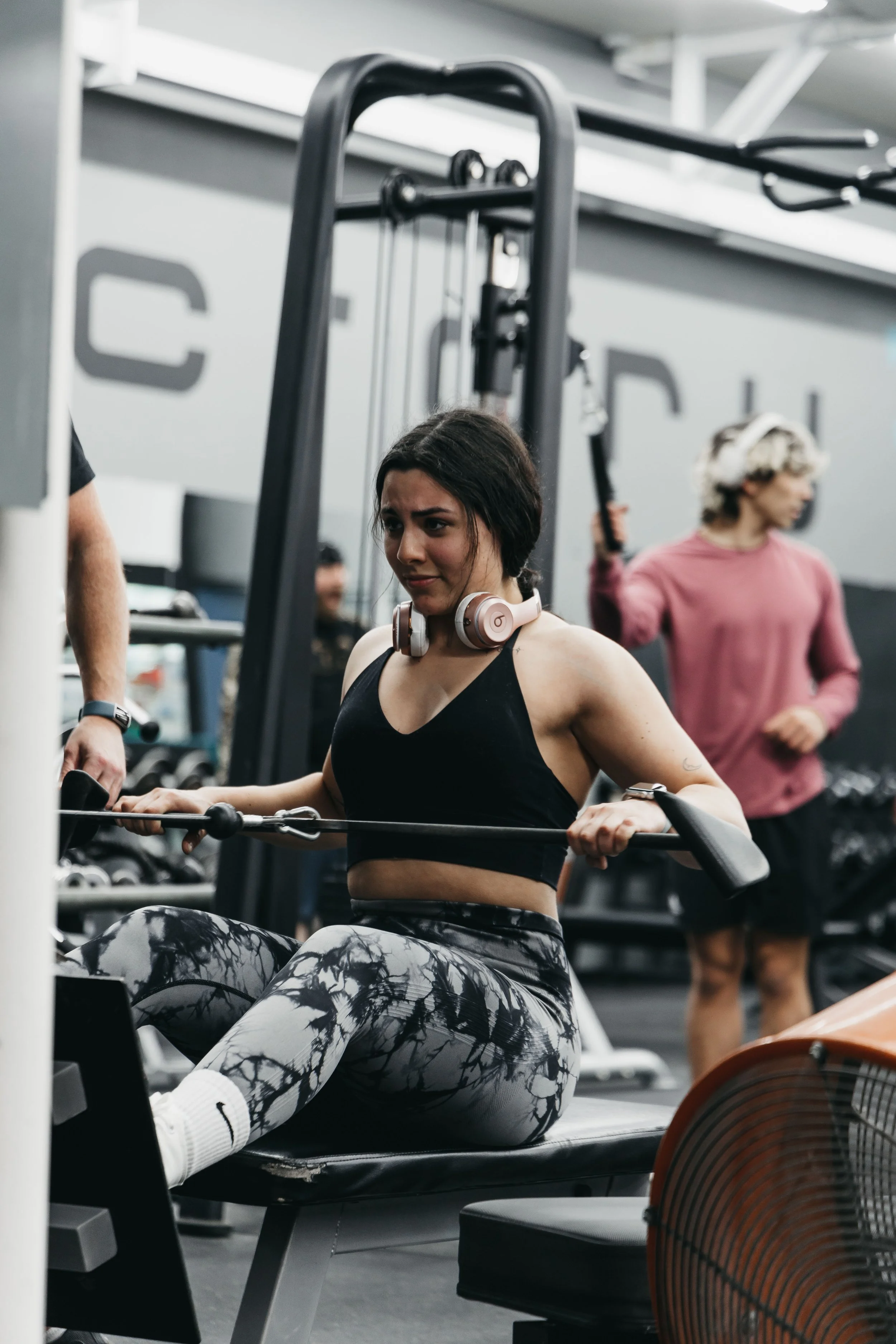 A woman in workout clothes with headphones around her neck is using a rowing machine at a gym, with other gym-goers in the background.
