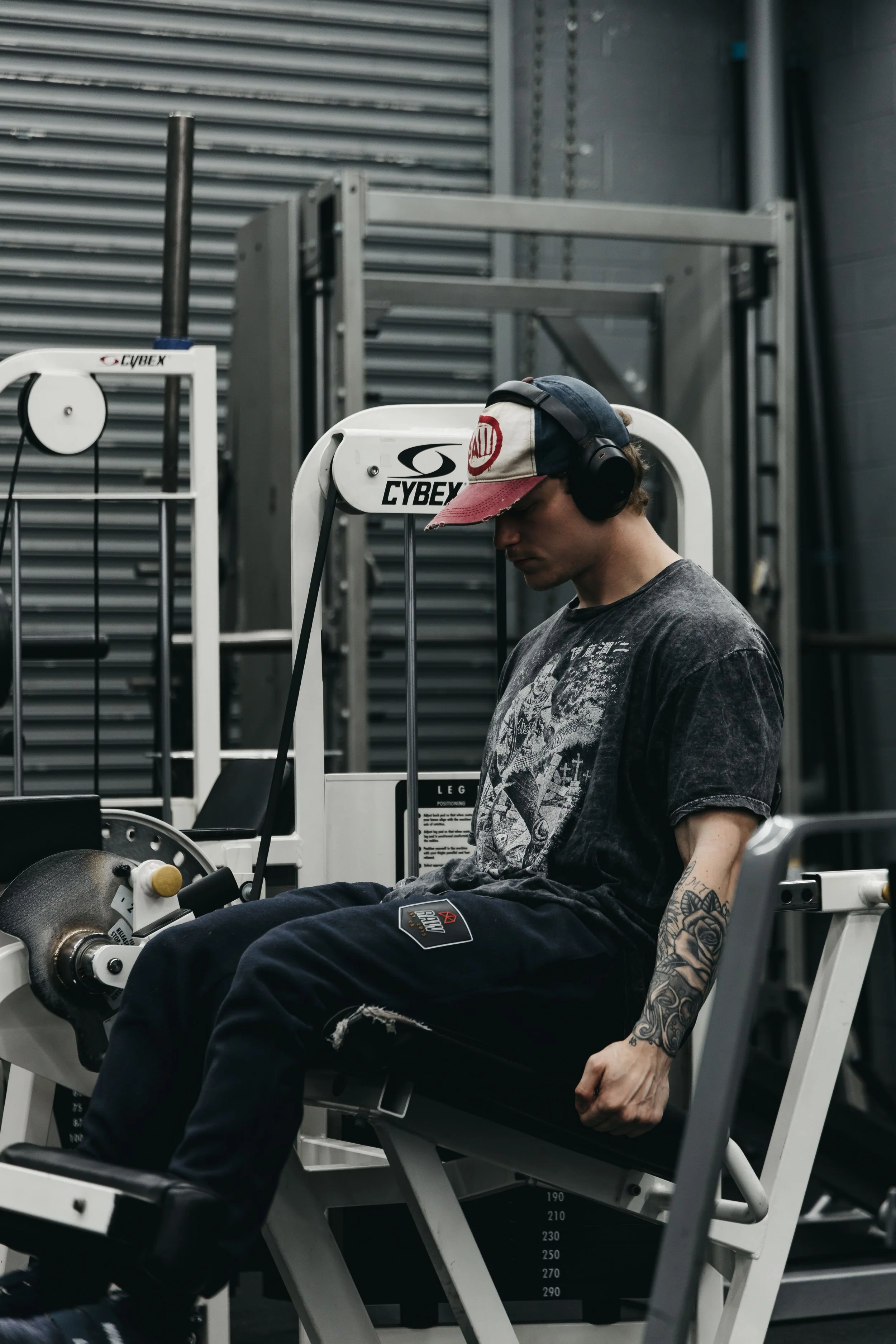 A young man with tattoos on his arm wearing a cap, headphones, and ripped jeans is performing leg extensions in a fitness center, focusing on his workout.