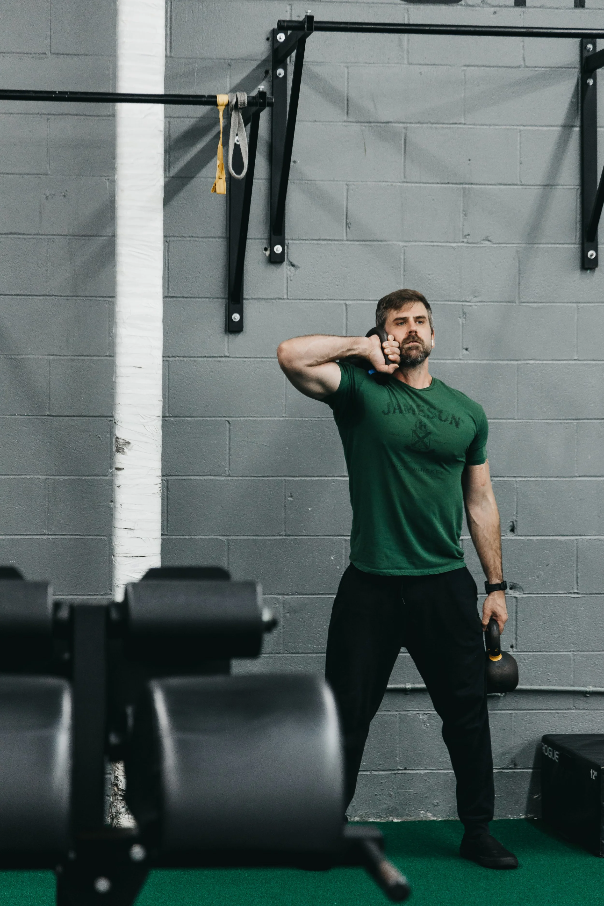 A man in a green T-shirt and black pants is standing in a gym, holding two kettlebells. He is performing a workout while standing on a green turff, with a gray brick wall and workout equipment in the background.