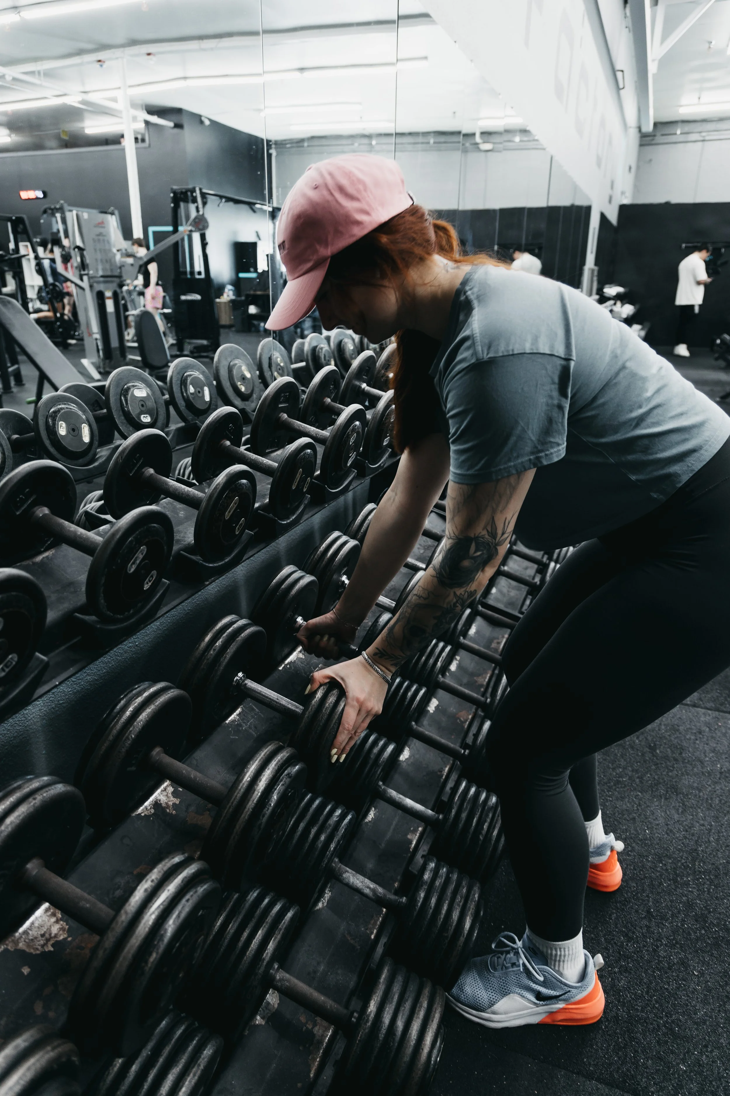 A woman wearing a pink baseball cap, a gray t-shirt, black leggings, gray sneakers with orange soles, and white socks, is bending over in front of a row of dumbbells at a gym, picking up weights.
