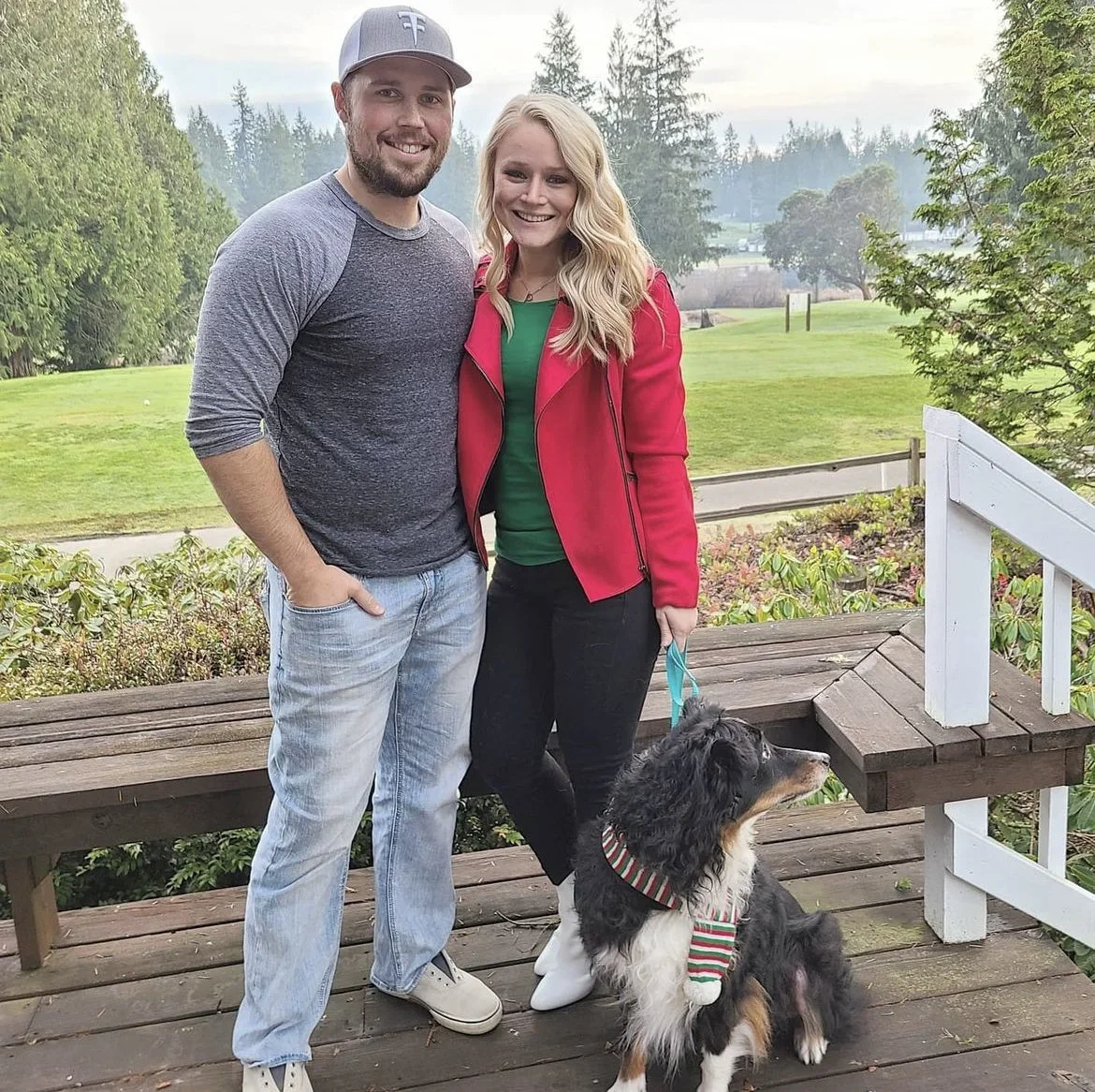 A smiling man and woman stand on a wooden deck with a dog, in a green park setting with trees and grass in the background.