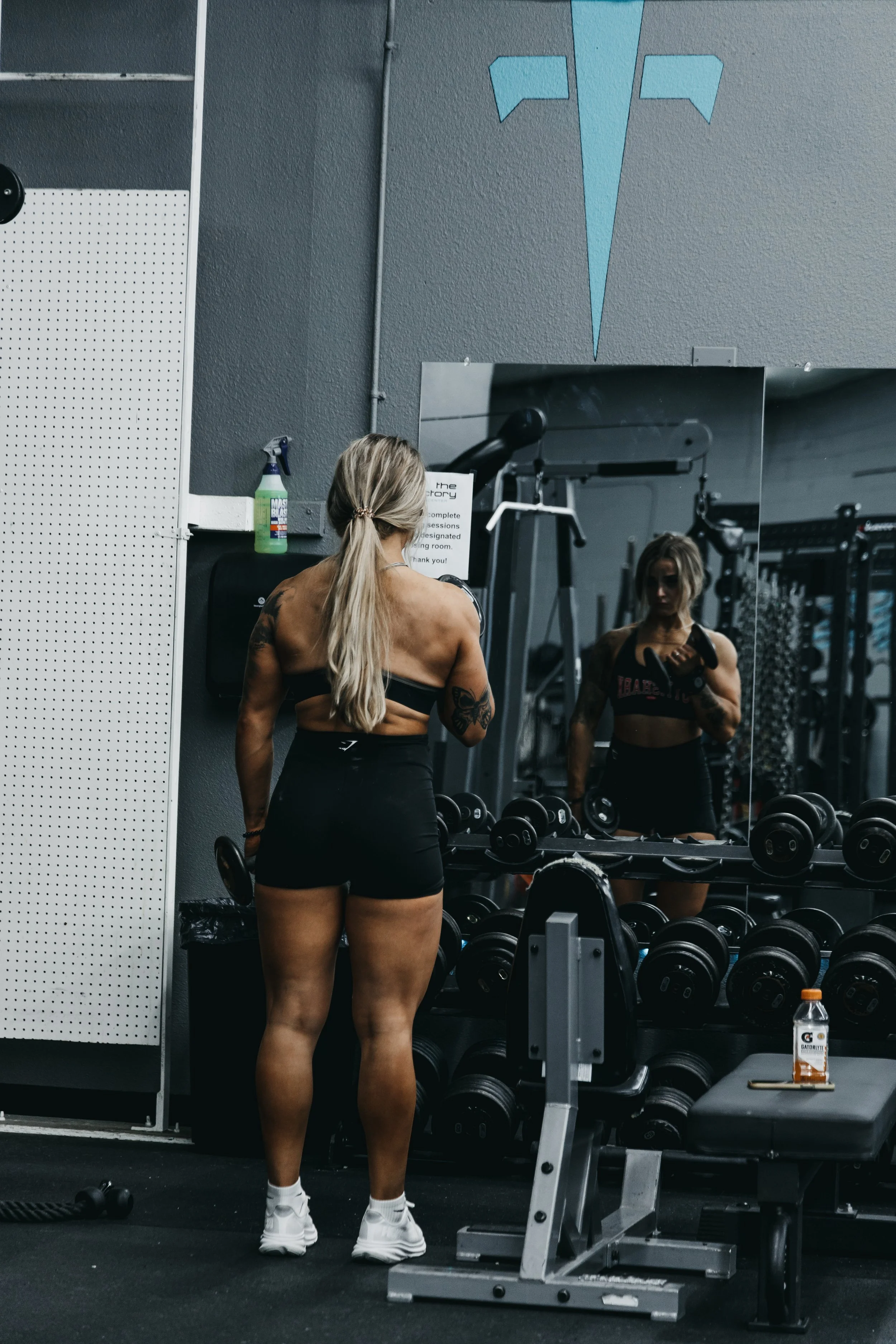 A woman in workout clothes standing in front of a mirror in a gym, with dumbbells on a rack, a water bottle on a bench, and fitness equipment in the background.
