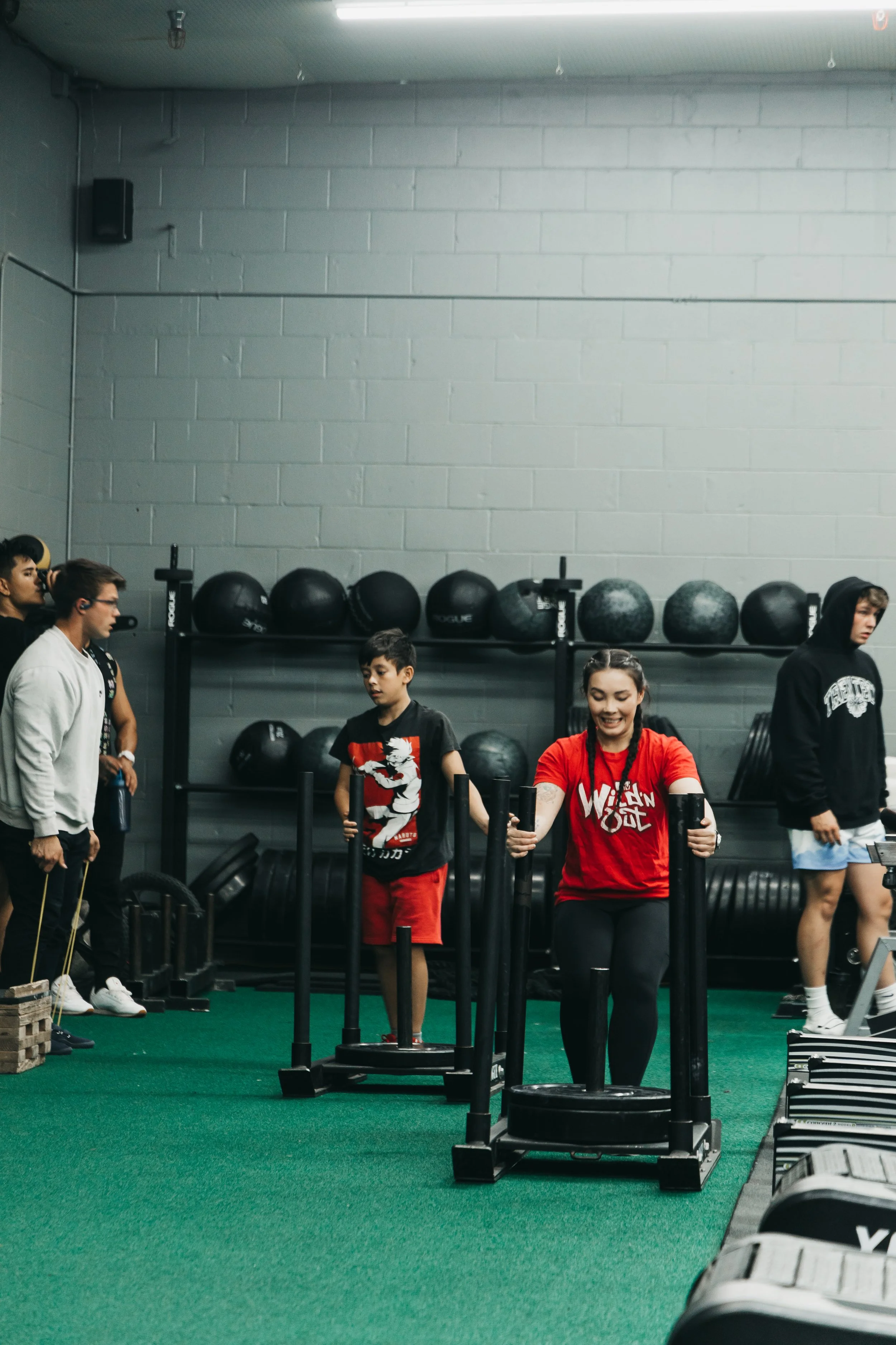 People working out in a gym with black medicine balls on a rack, green turf flooring, and gray cinder block walls.