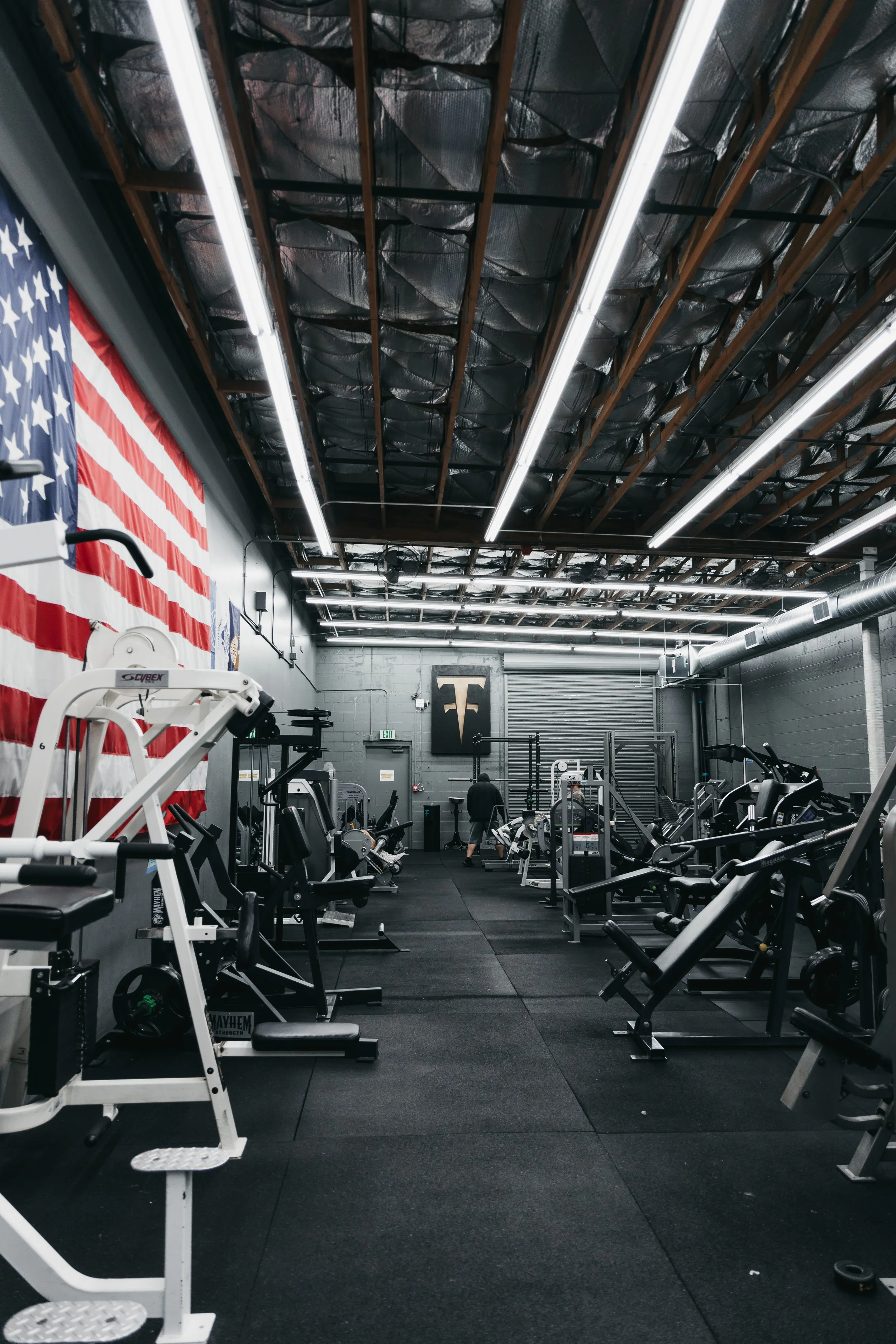 Interior of a gym with fitness equipment, a large American flag on the wall, and a logo on the wall, with high ceilings and industrial lighting.