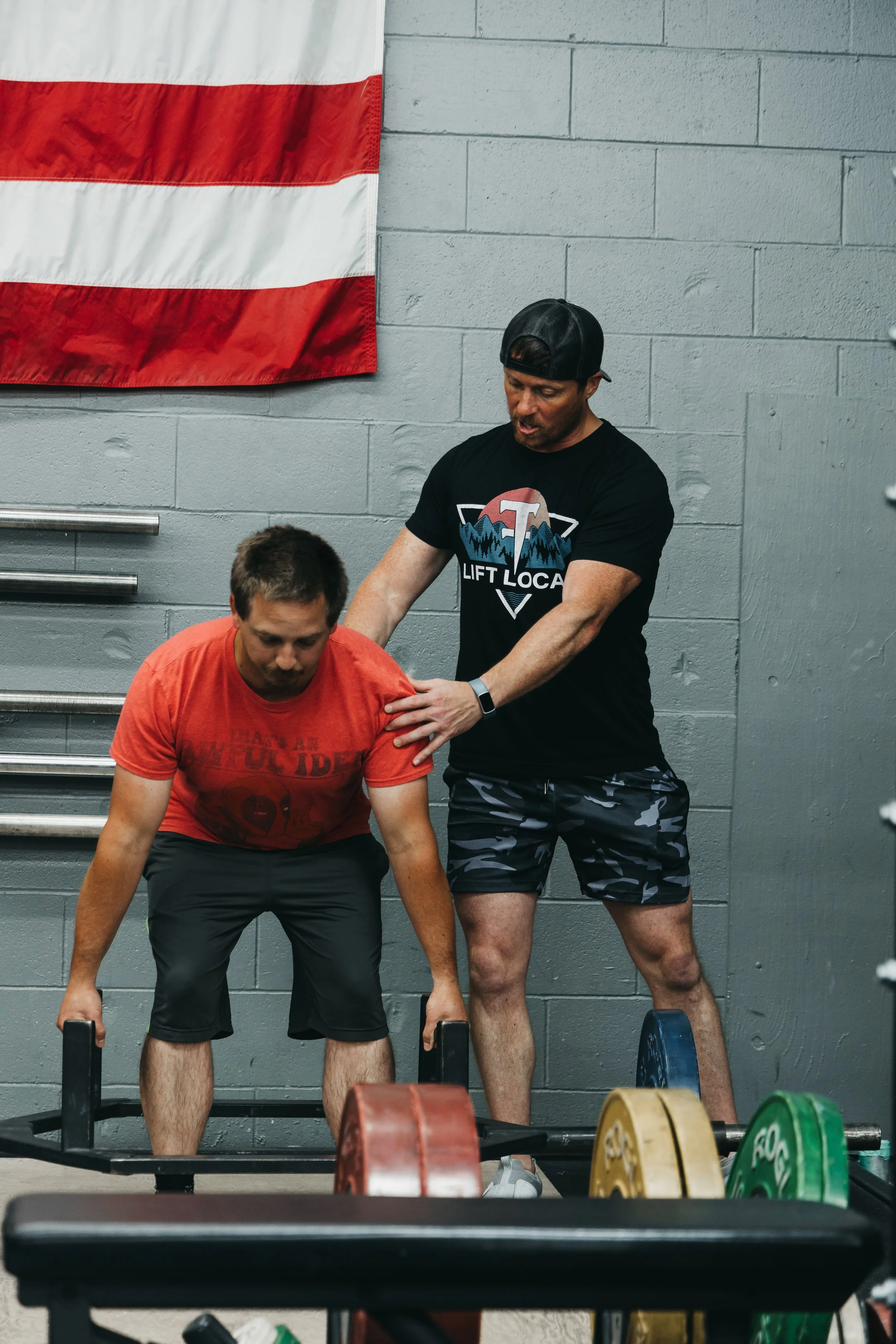 Two men in a gym performing deadlift exercises with weight plates on a barbell, with one man assisting the other.