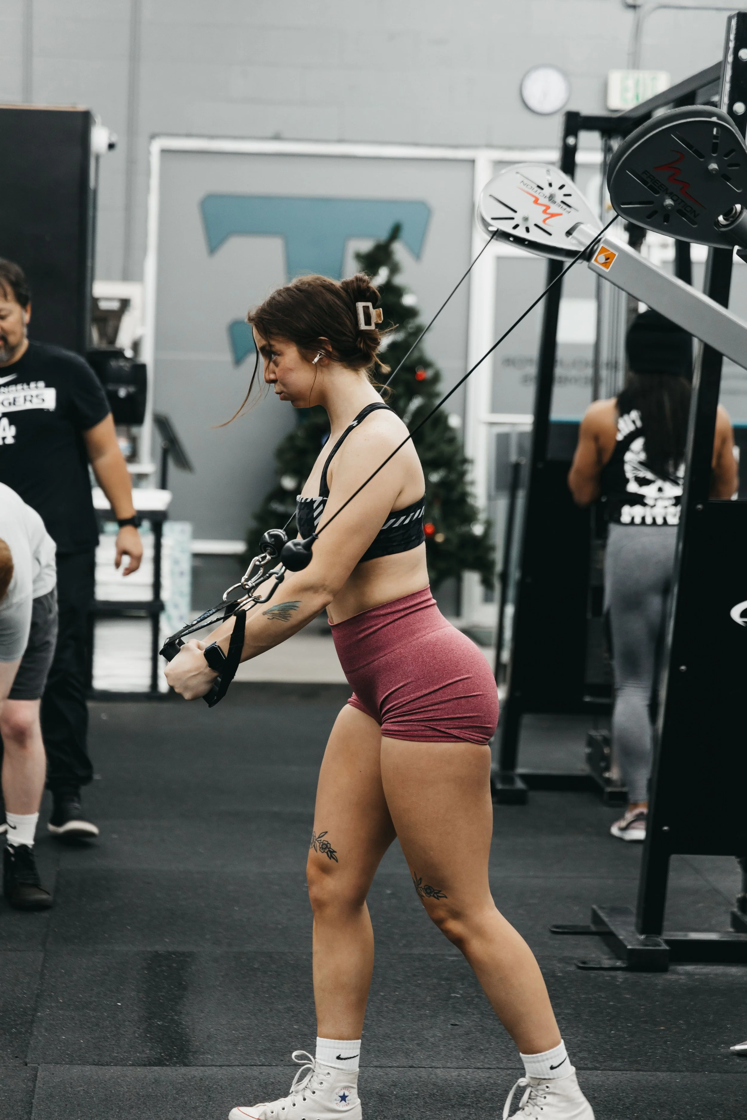 A woman in workout attire is using a cable machine at the gym, with weights and exercise equipment visible around her, and a Christmas tree in the background.