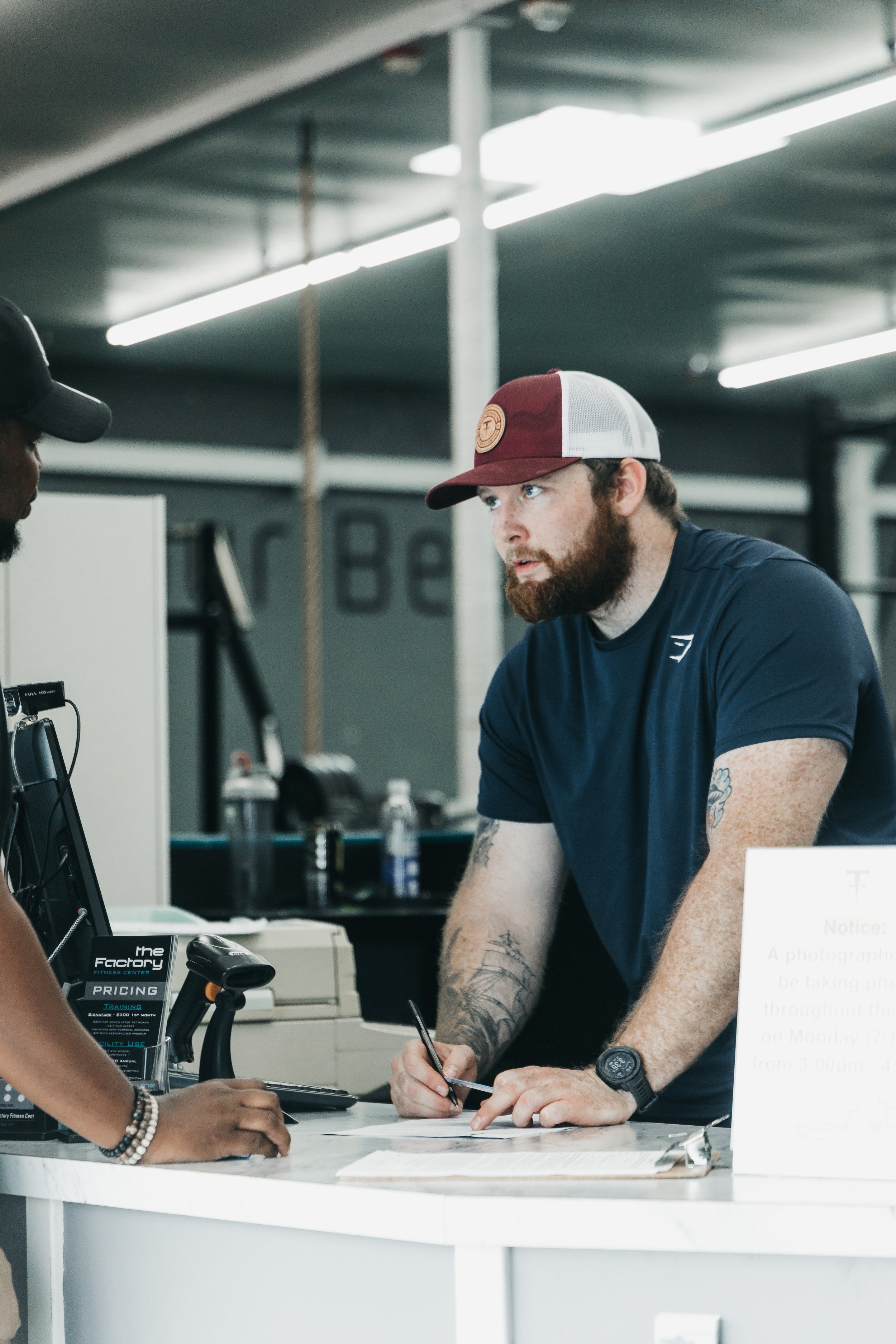 A man with a beard and tattoos is signing a document at a counter, while a woman hands him a payment card. The background suggests a modern, industrial-style setting, possibly a fitness center or similar establishment.
