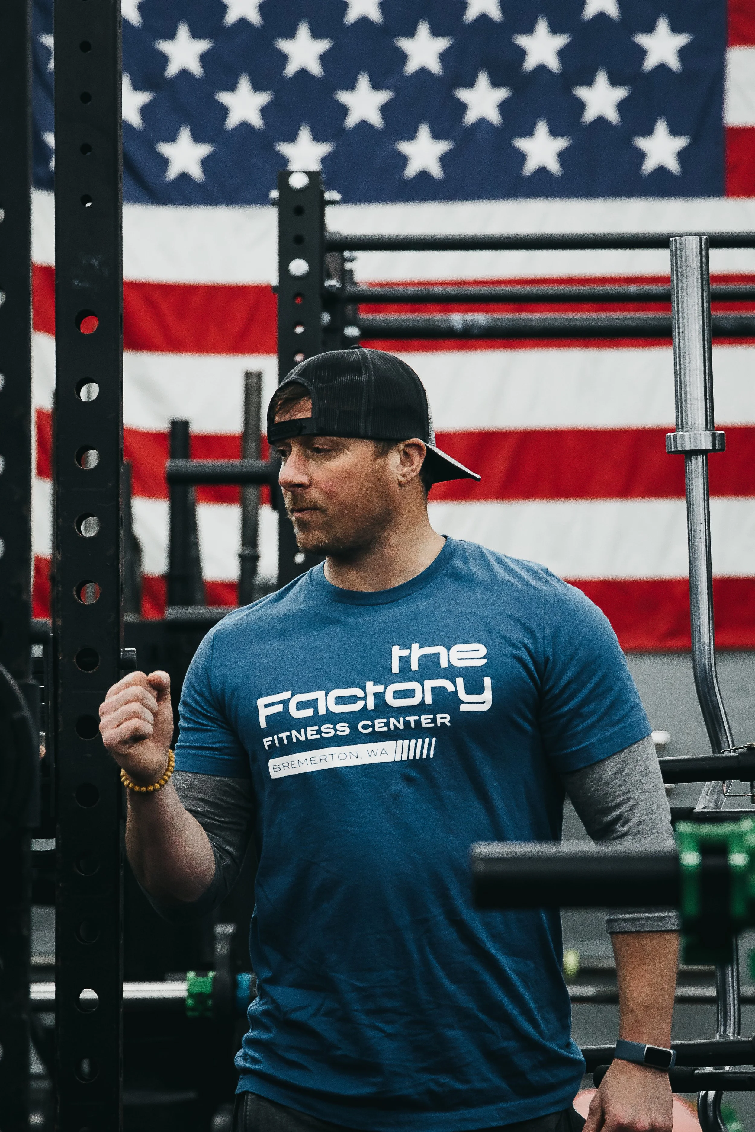 A man in a blue T-shirt and a black backward cap standing in a gym, with an American flag hanging on the wall behind him.