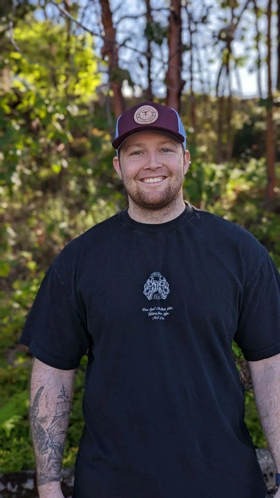 A smiling man with a beard and tattoos on his right arm, wearing a black T-shirt and a cap, standing outdoors in a green wooded area.