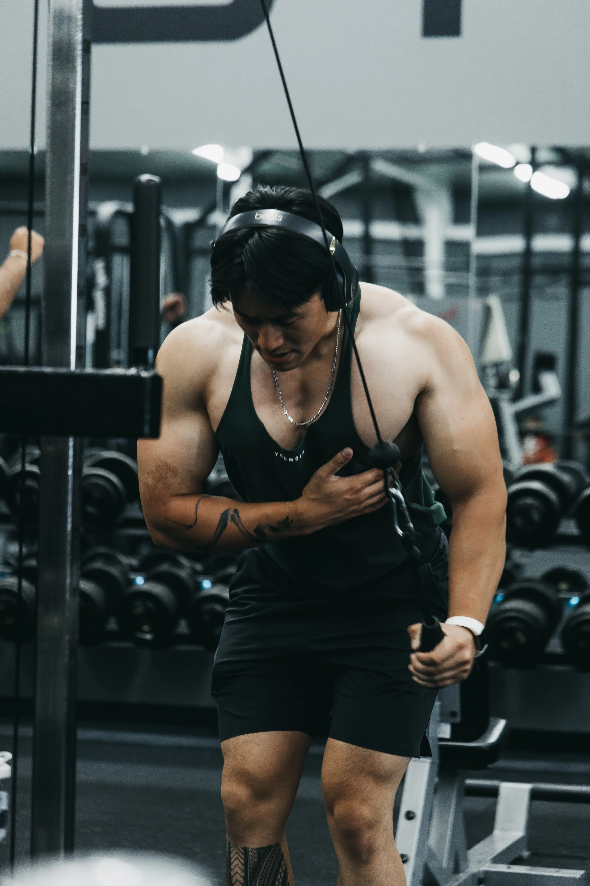 A muscular man with headphones, wearing a black tank top and shorts, is leaning on a gym machine with one hand on his chest, in a gym with dumbbells in the background.
