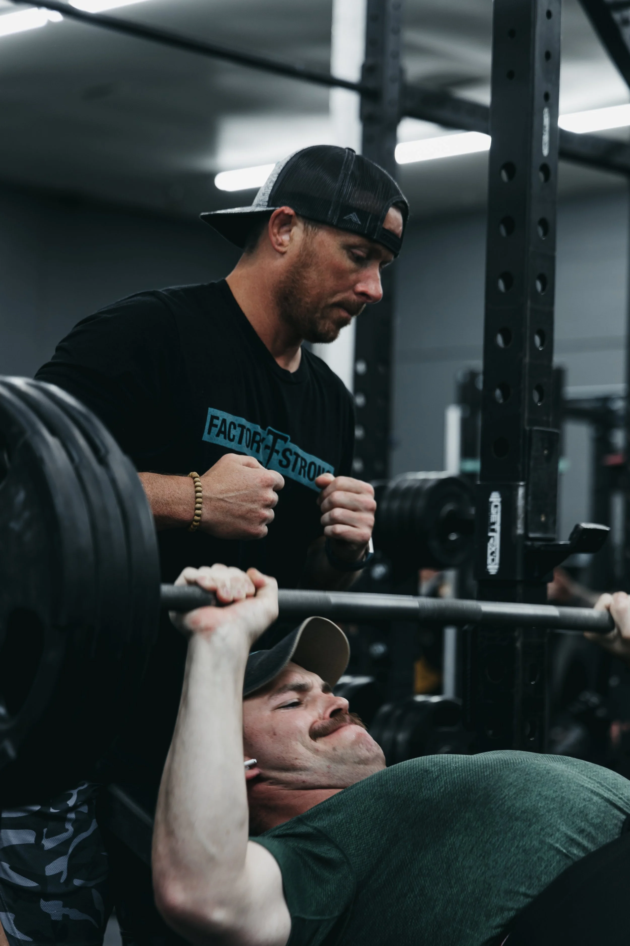 A man lying on a bench pressing a barbell while another man spots him in a gym.