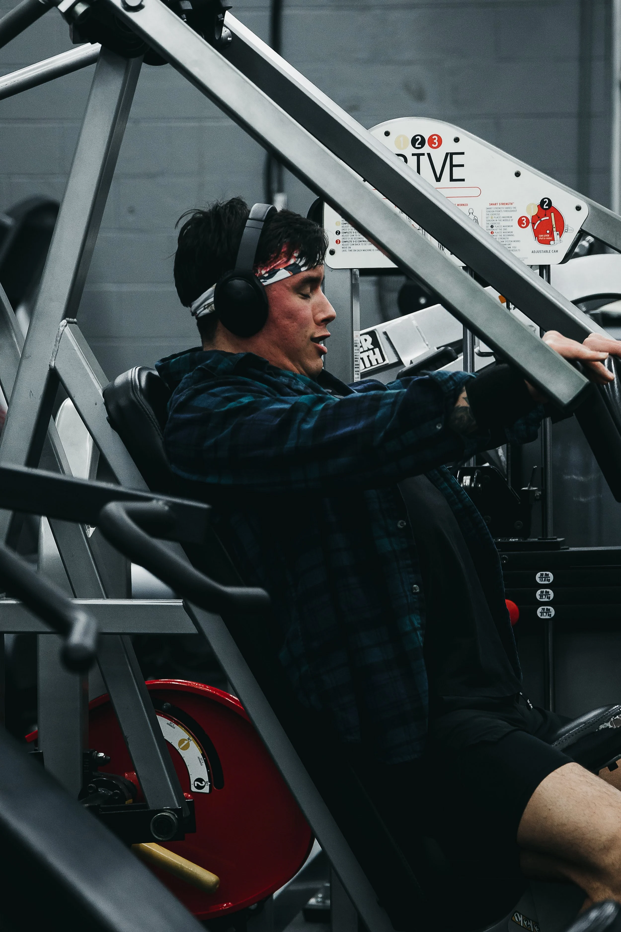 Young man with headphones performing a chest press on an exercise machine, eyes closed, in workout clothes, in a fitness gym.