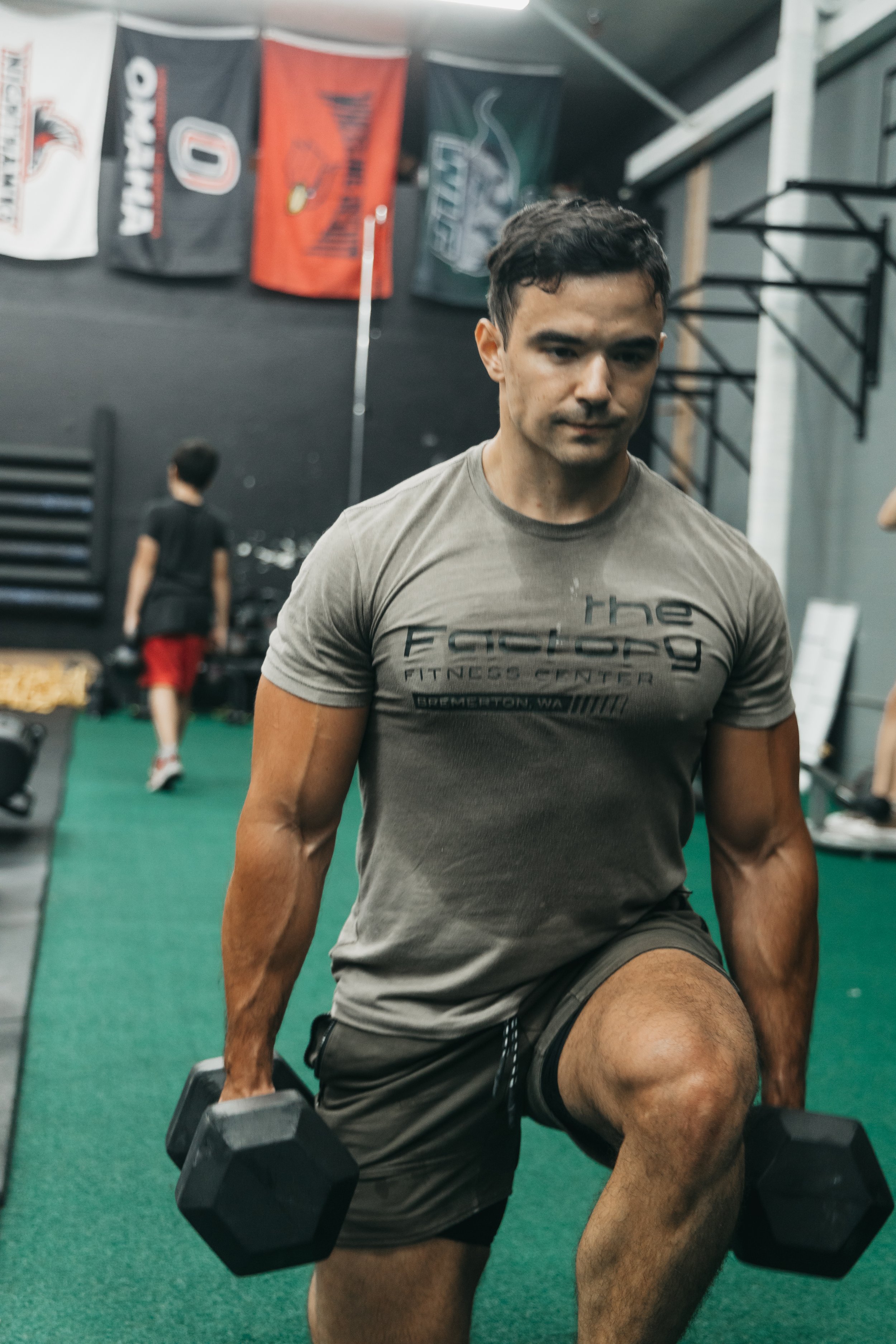A man in a gray t-shirt with black shorts performing a lunge with dumbbells in each hand in a gym. In the background, there are flags hanging on the wall and a person wearing a black shirt and red shorts walking away.