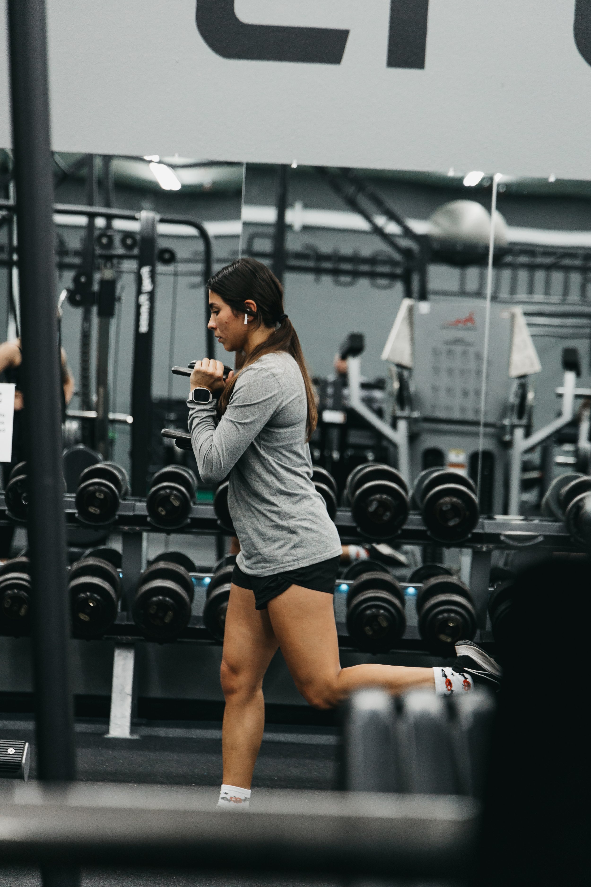 A woman doing a Bulgarian split squat exercise in a gym, surrounded by weights and gym equipment.