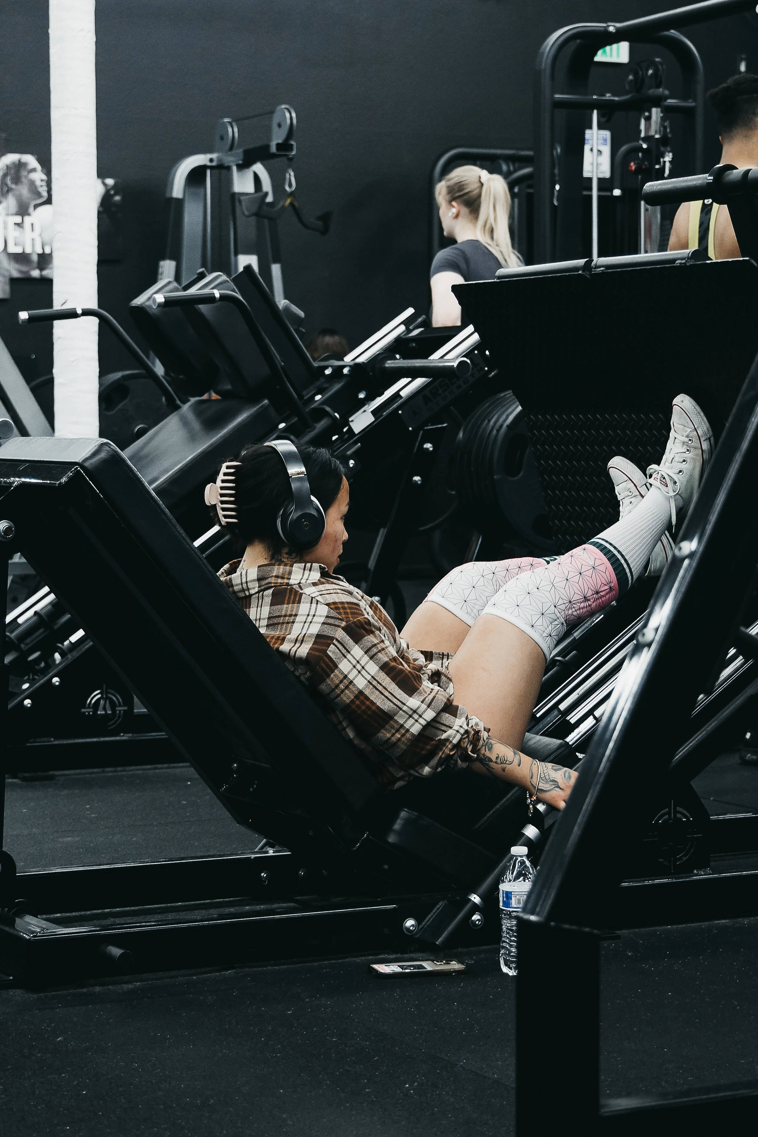 A woman with tattoos and headphones sitting on a leg press machine with her legs raised, wearing a plaid shirt, shorts, and sneakers in a gym. Other gym equipment and people working out are in the background.