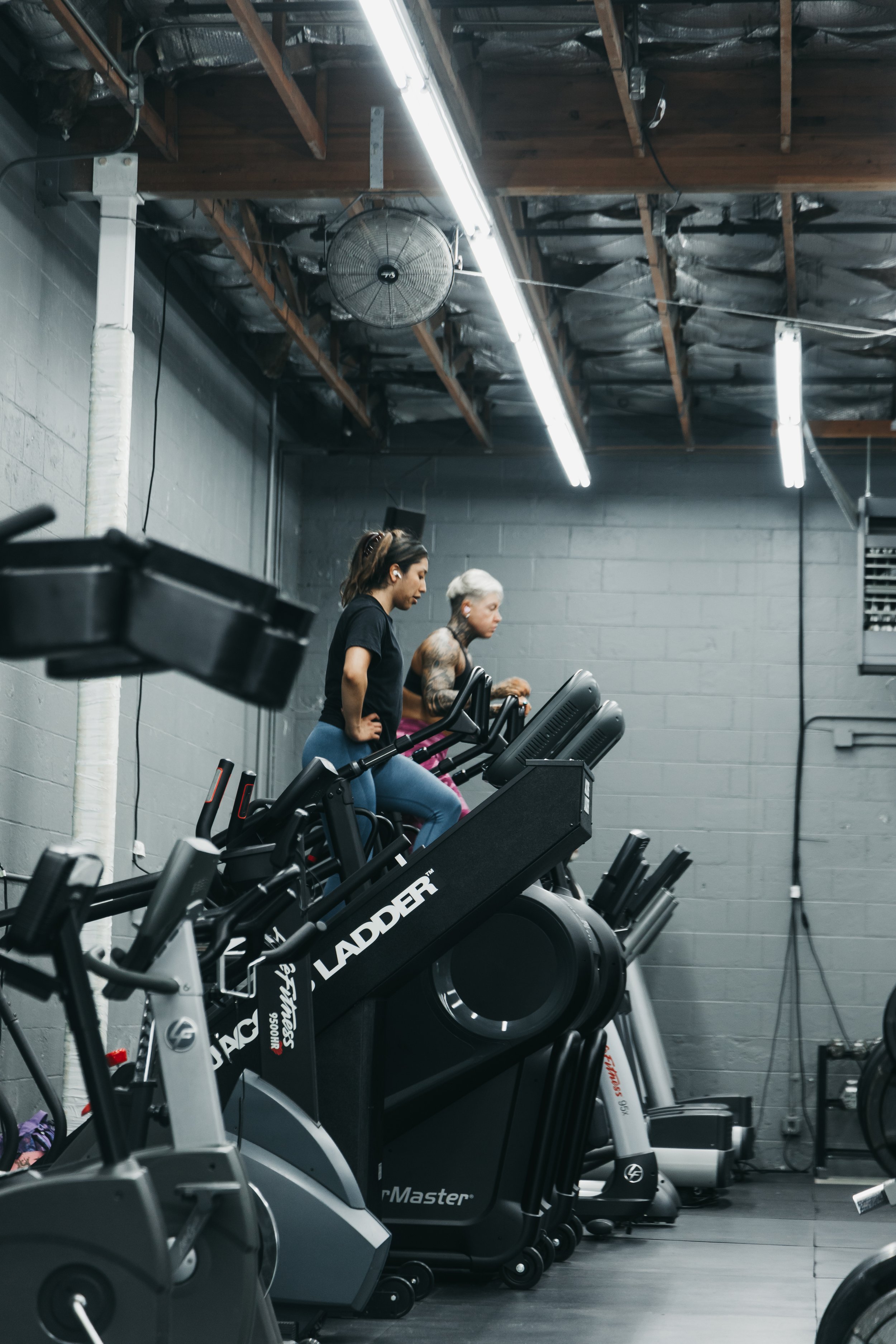 Two women on Stairmasters in a gym, with industrial ceiling and fluorescent lighting.