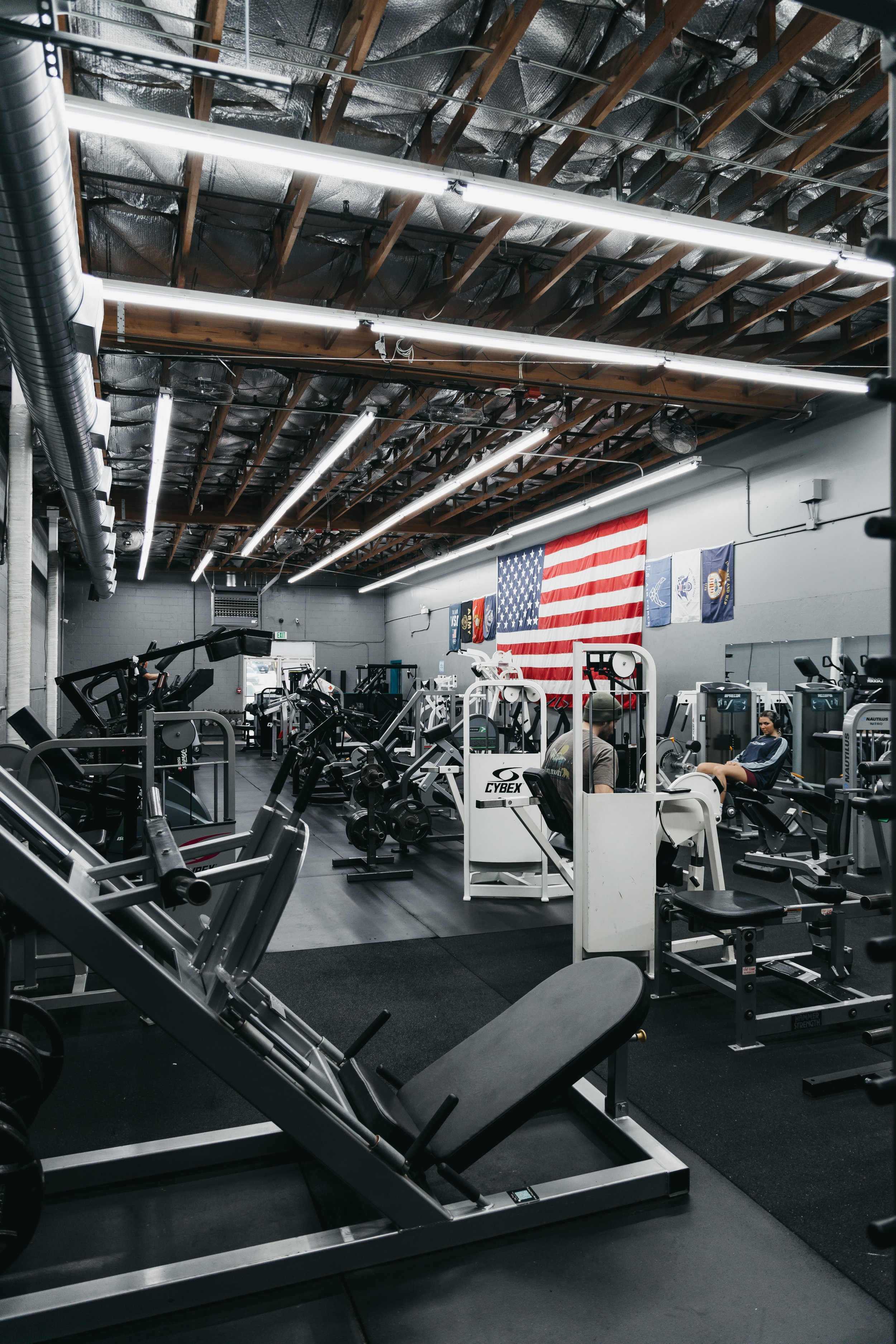 Inside a gym with various exercise equipment, including treadmills and weight machines, with American flags hanging on the wall and fluorescent lighting on the ceiling.
