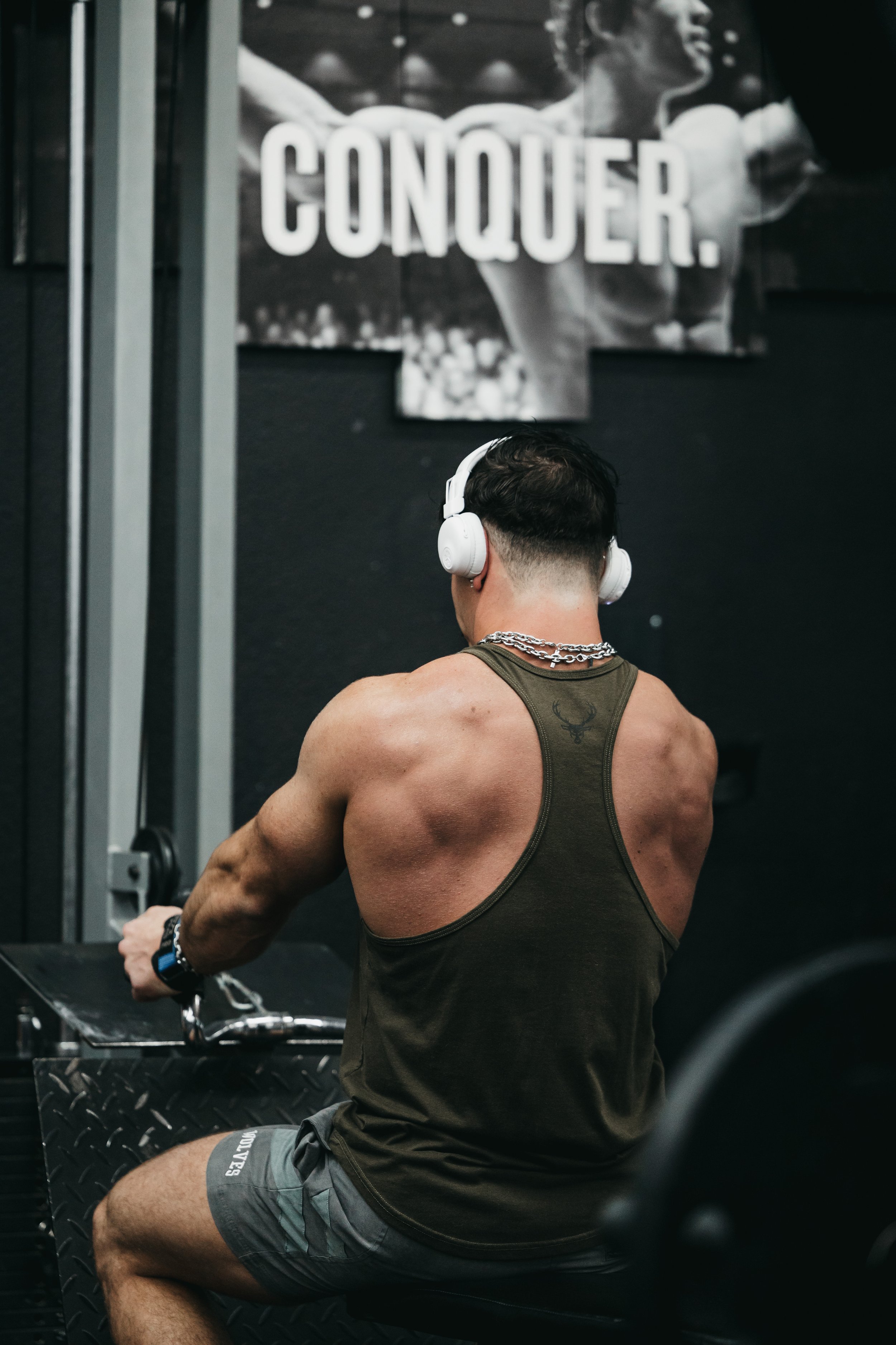 A man working out on a rowing machine in a gym. He is wearing headphones, a green tank top, gray shorts, and a chain necklace. Behind him is a black and white poster with the word 'Conquer' and Arnold Schwarzenegger.