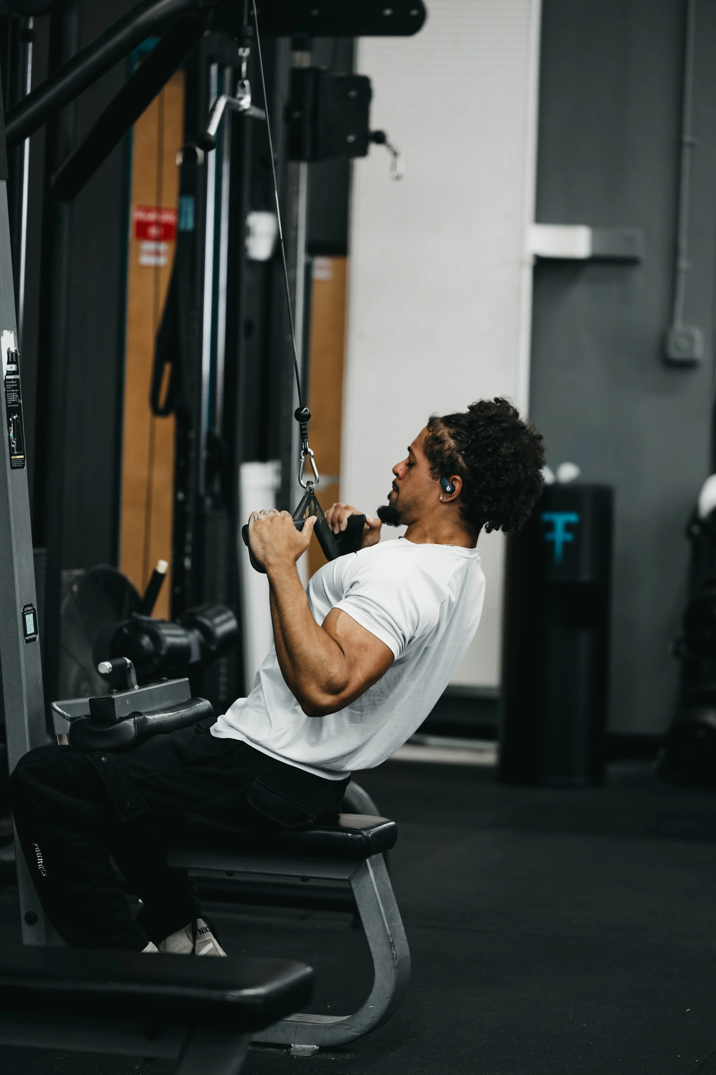 A man with curly hair and earbuds performing a lat pulldown exercise in a gym.