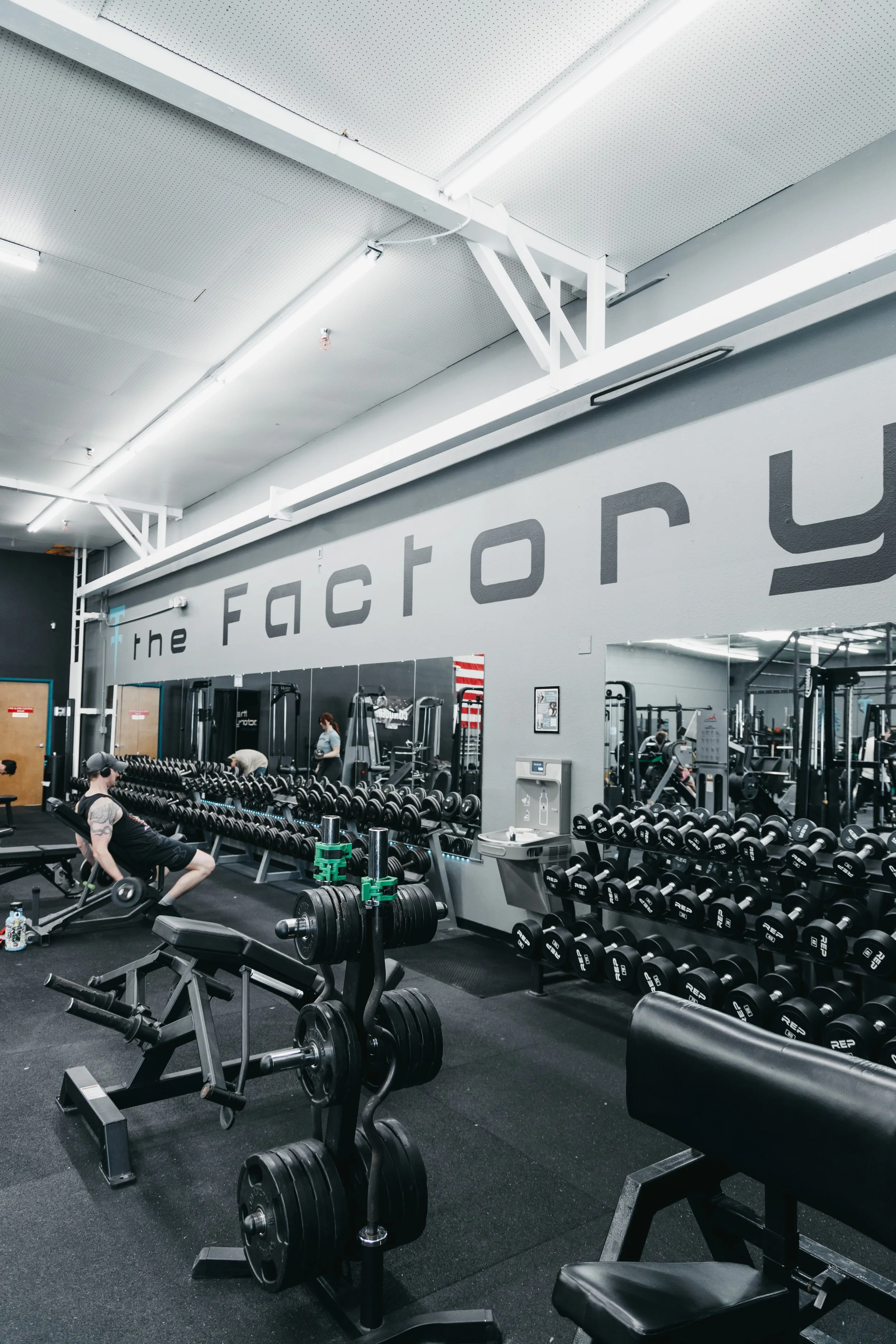 Interior of a gym with various weightlifting equipment, including free weights, dumbbells, and benches, and people working out.