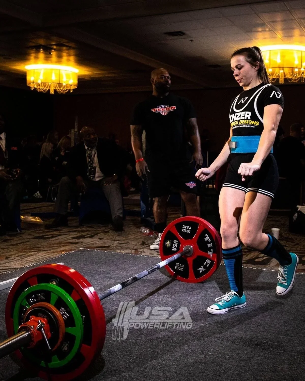 A woman in athletic clothing performing a deadlift with a barbell at a powerlifting event, with a judge watching and an audience seated in the background.