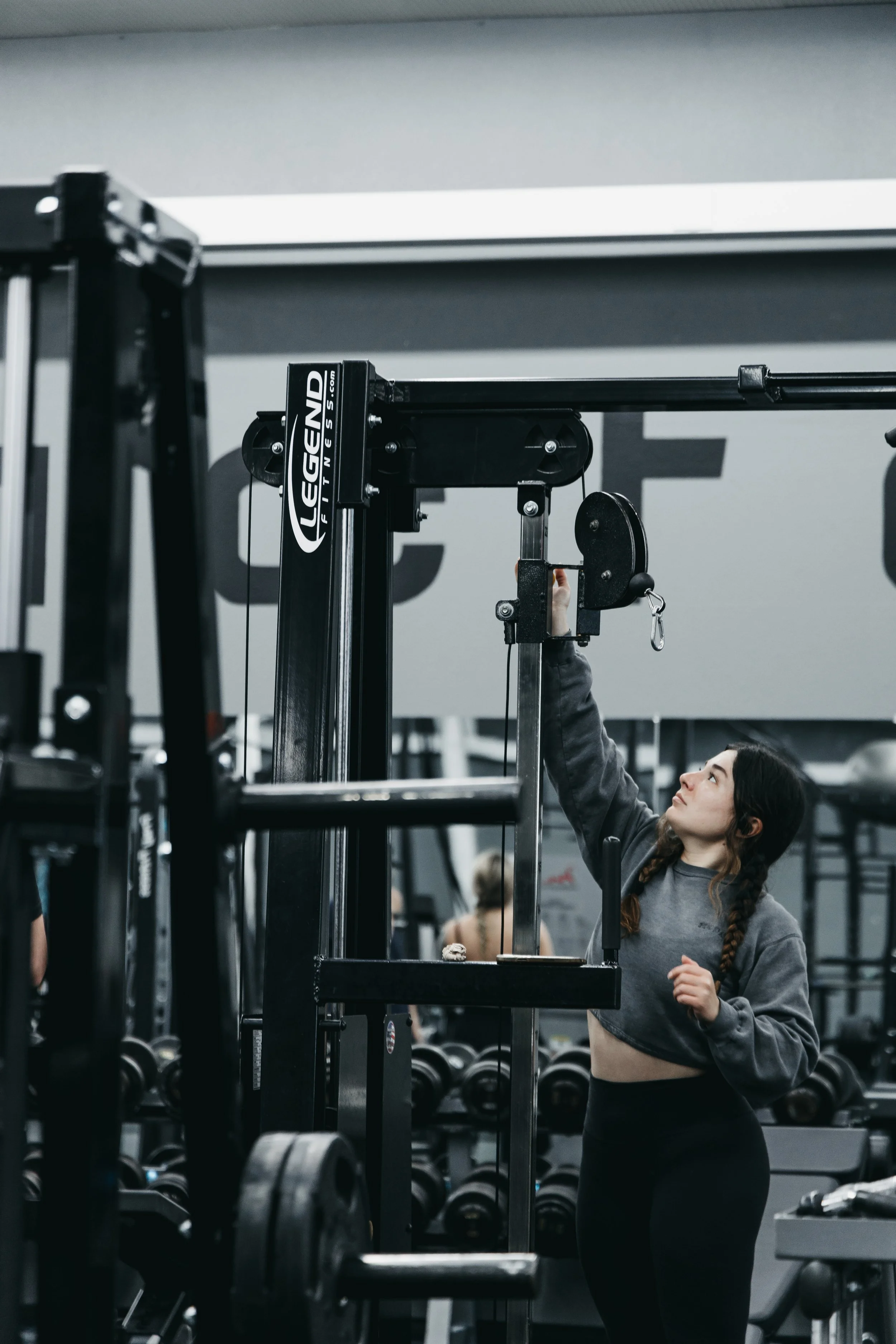 A woman in a gym is adjusting a cable weight machine. The gym appears to have multiple weights and exercise equipment. She has long braided hair and is wearing a gray hoodie.