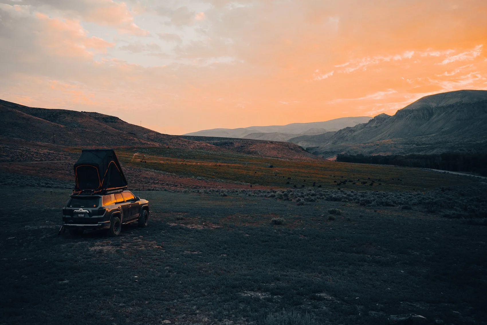 A black SUV with a rooftop tent parked on a grassy field in a mountainous landscape during sunset, with a sky painted in soft orange and pink hues.