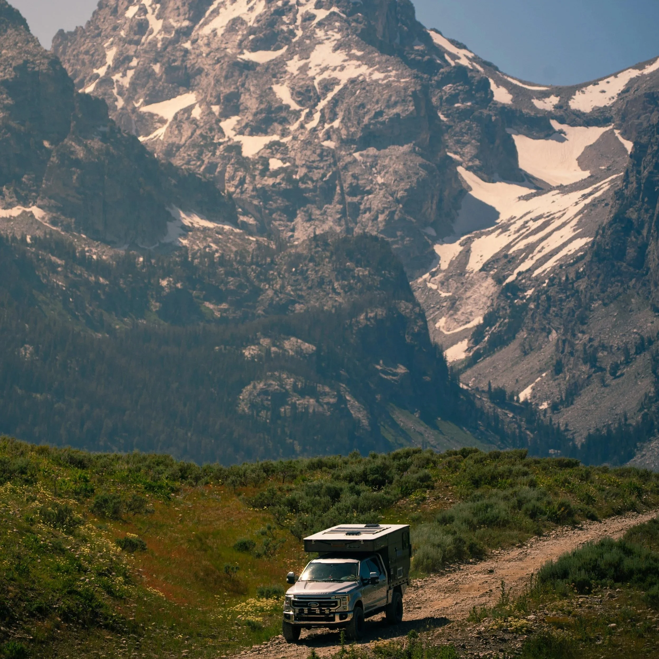 A rugged mountain range, the Tetons,  with snow patches and some green vegetation at the base. In the foreground, a truck with a camper is parked on a dirt trail in a remote, scenic wilderness area.