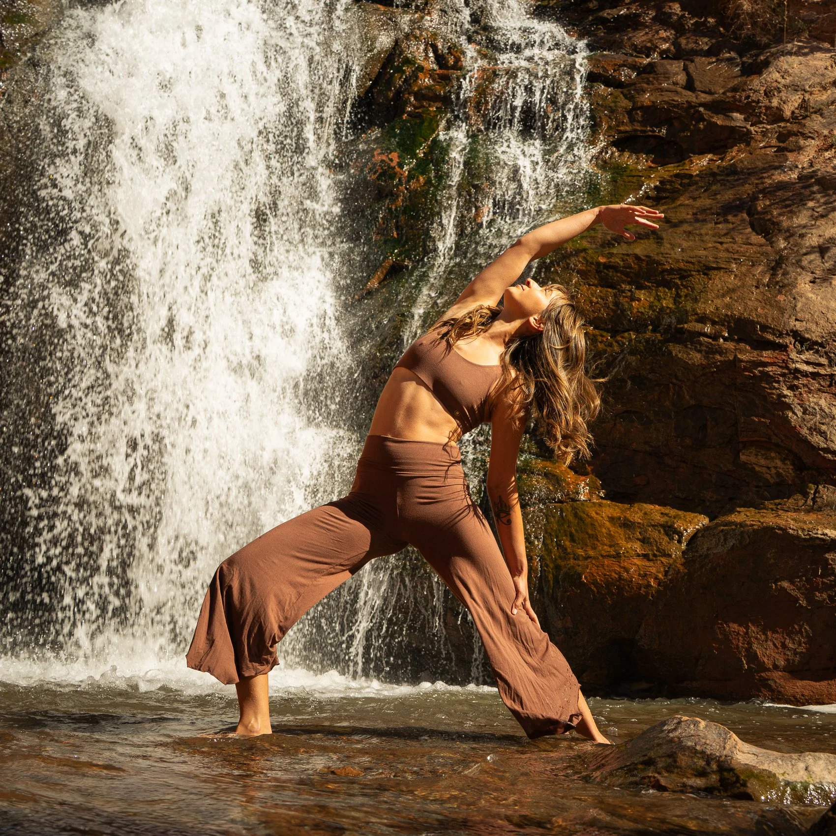 A woman with wet hair and brown clothing dancing in front of a waterfall and rocks, with water splashing at her feet.