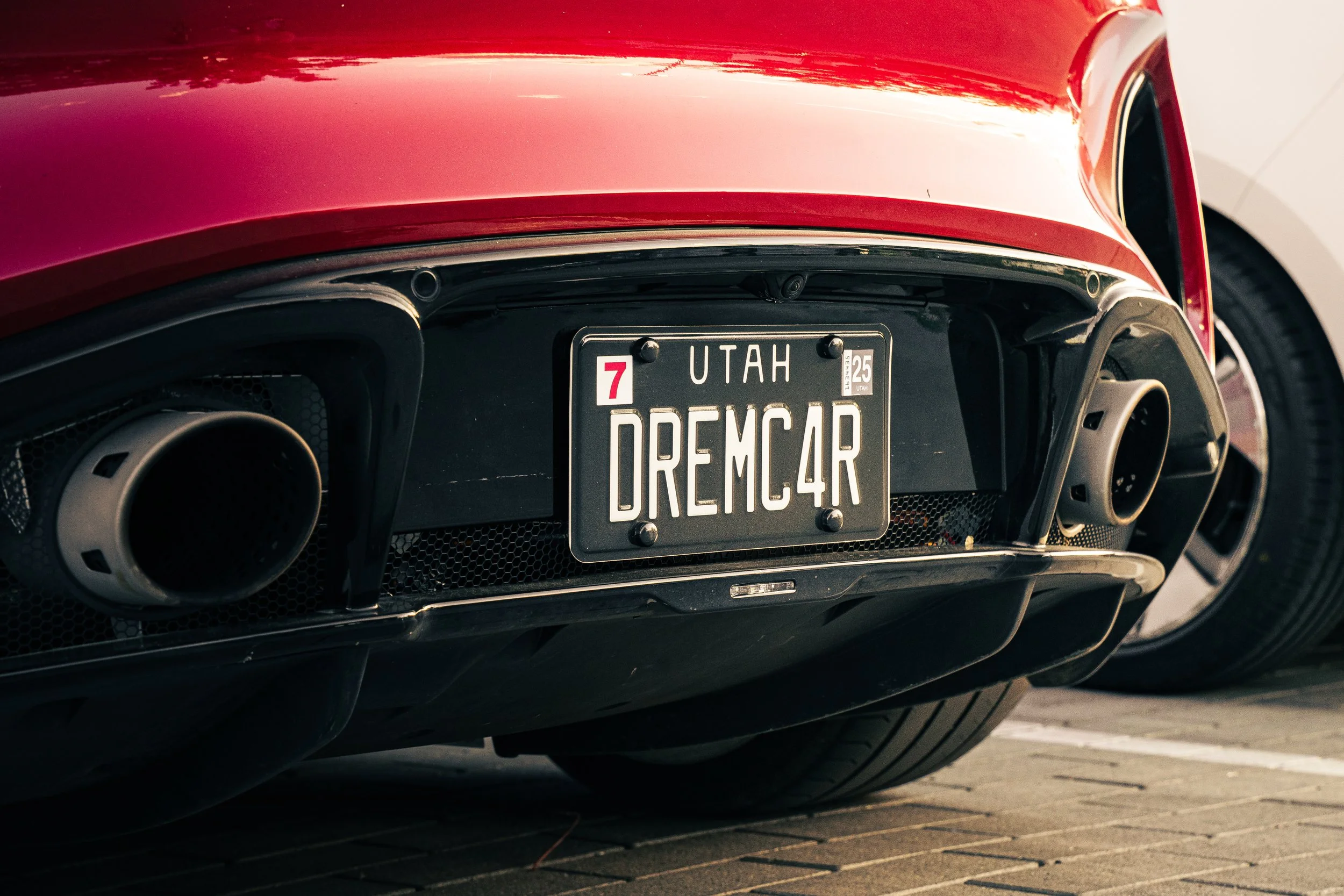 Close-up of the rear end of a red sports car, showing the exhaust pipes, dark rear bumper, and a custom Utah license plate reading "DREMC4R" with a red number 7 and a small number 25 in the top right corner.