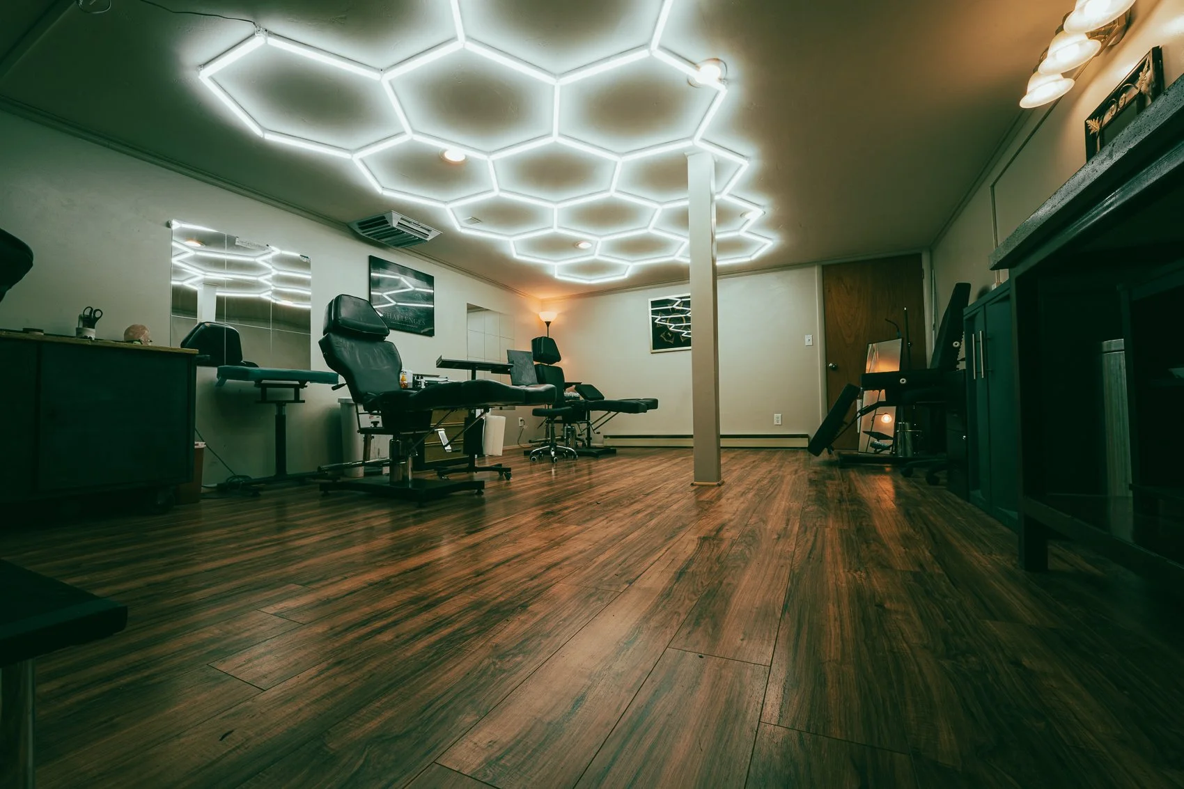 Empty wellness or tattoo clinic room with massage chairs, mirrors, and exercise equipment. Wooden floor, modern lighting, and neutral walls.