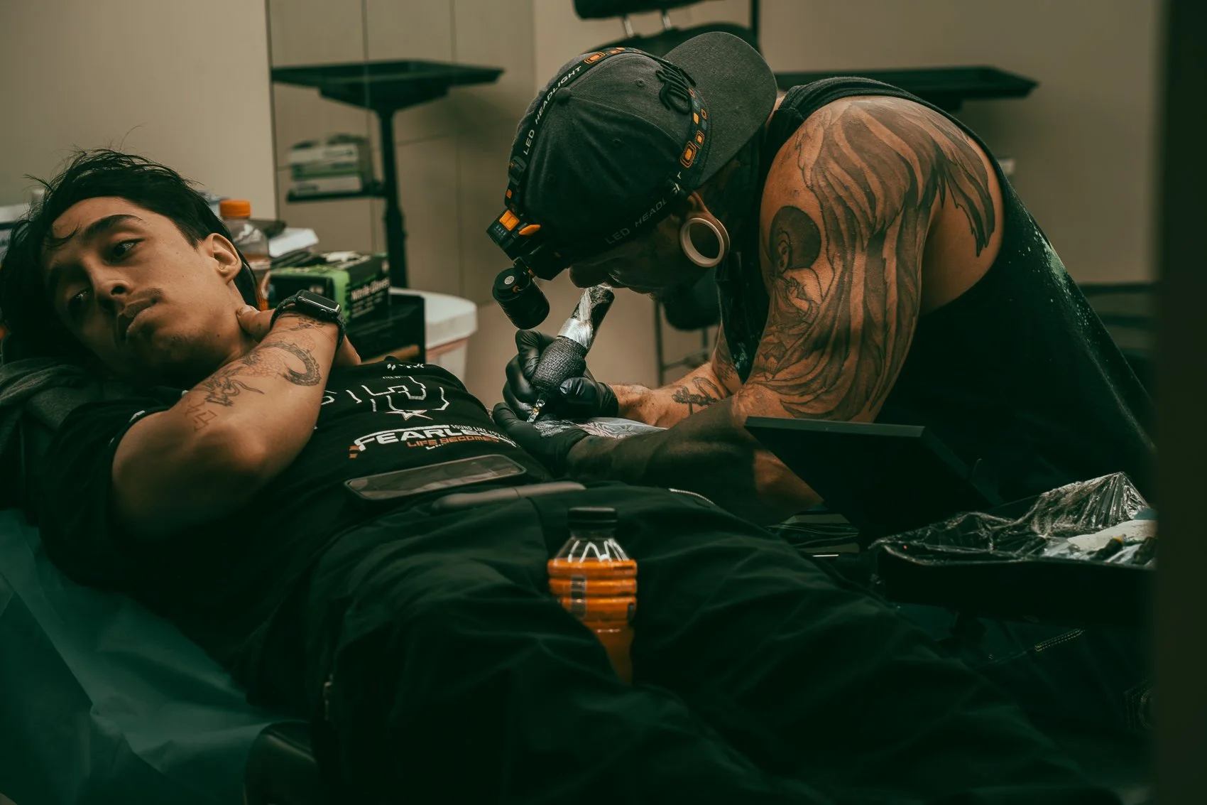 A man with tattoos lying on a bed while a tattoo artist with tattoos is working on his arm in a tattoo studio.